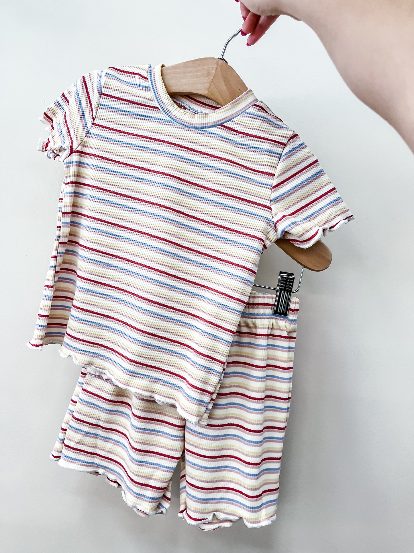 A hand holds a wooden hanger displaying the Sweet Stripe Snuggle Set by Tiny Tot Bundles: a matching children’s short-sleeve ribbed top and shorts featuring colorful horizontal stripes on white.