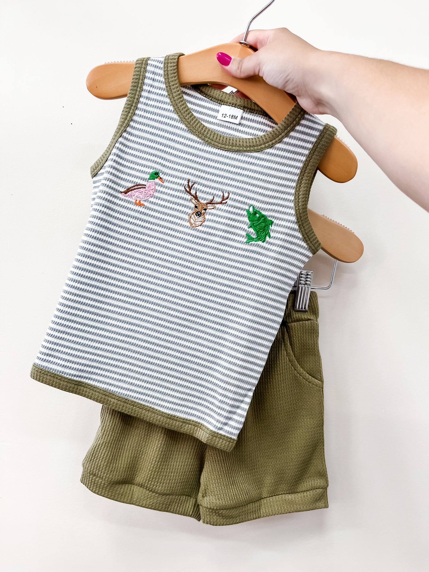 A hand displays the Tiny Tot Bundles Woodland Adventure Tank Set—a sleeveless striped top with animal embroidery and matching green shorts—on a hanger.
