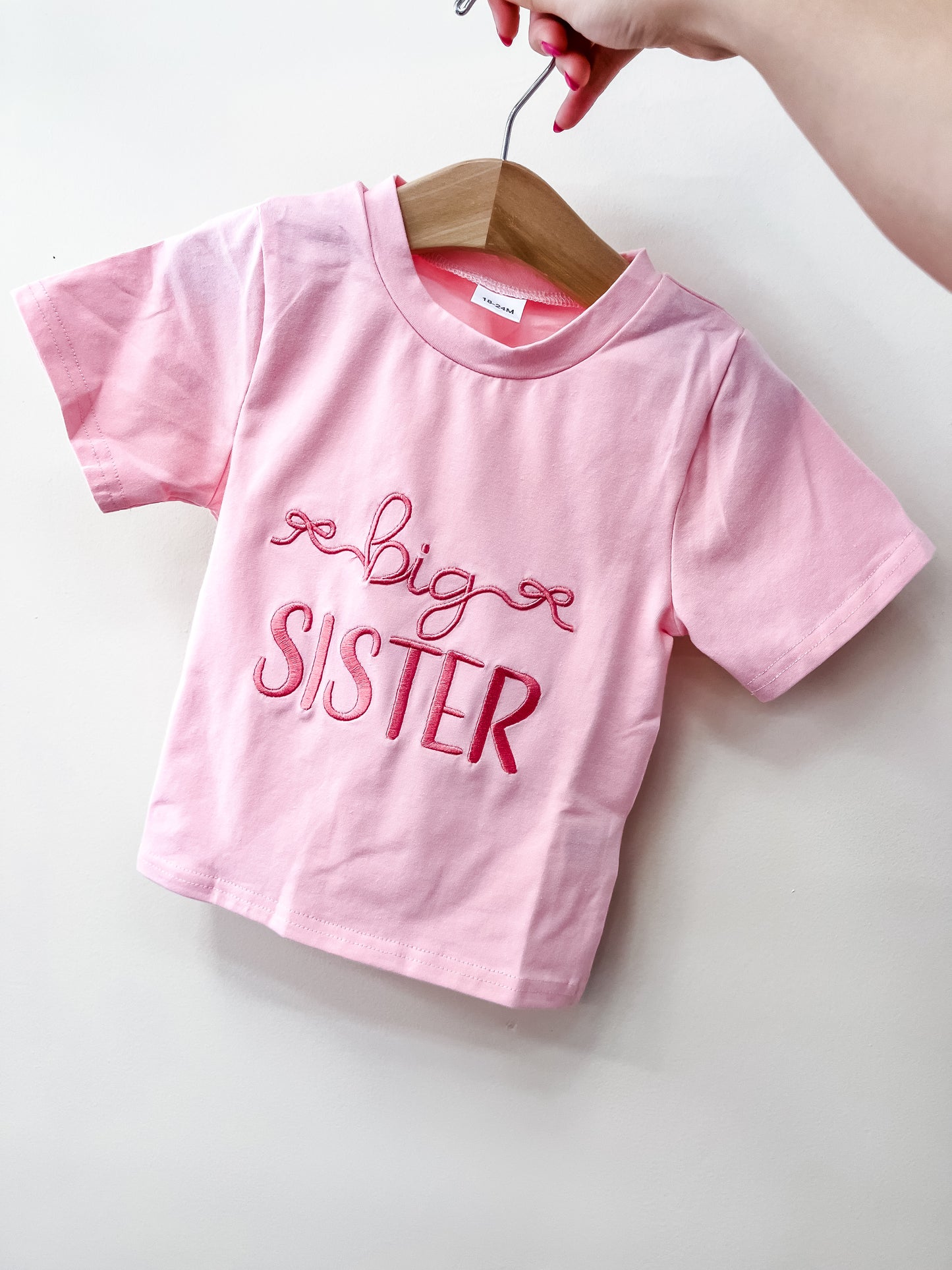 A hand holds a hanger with a pink Tiny Tot Bundles Big Sister Bow T-Shirt featuring "big SISTER" embroidered in red and pink script on the front.