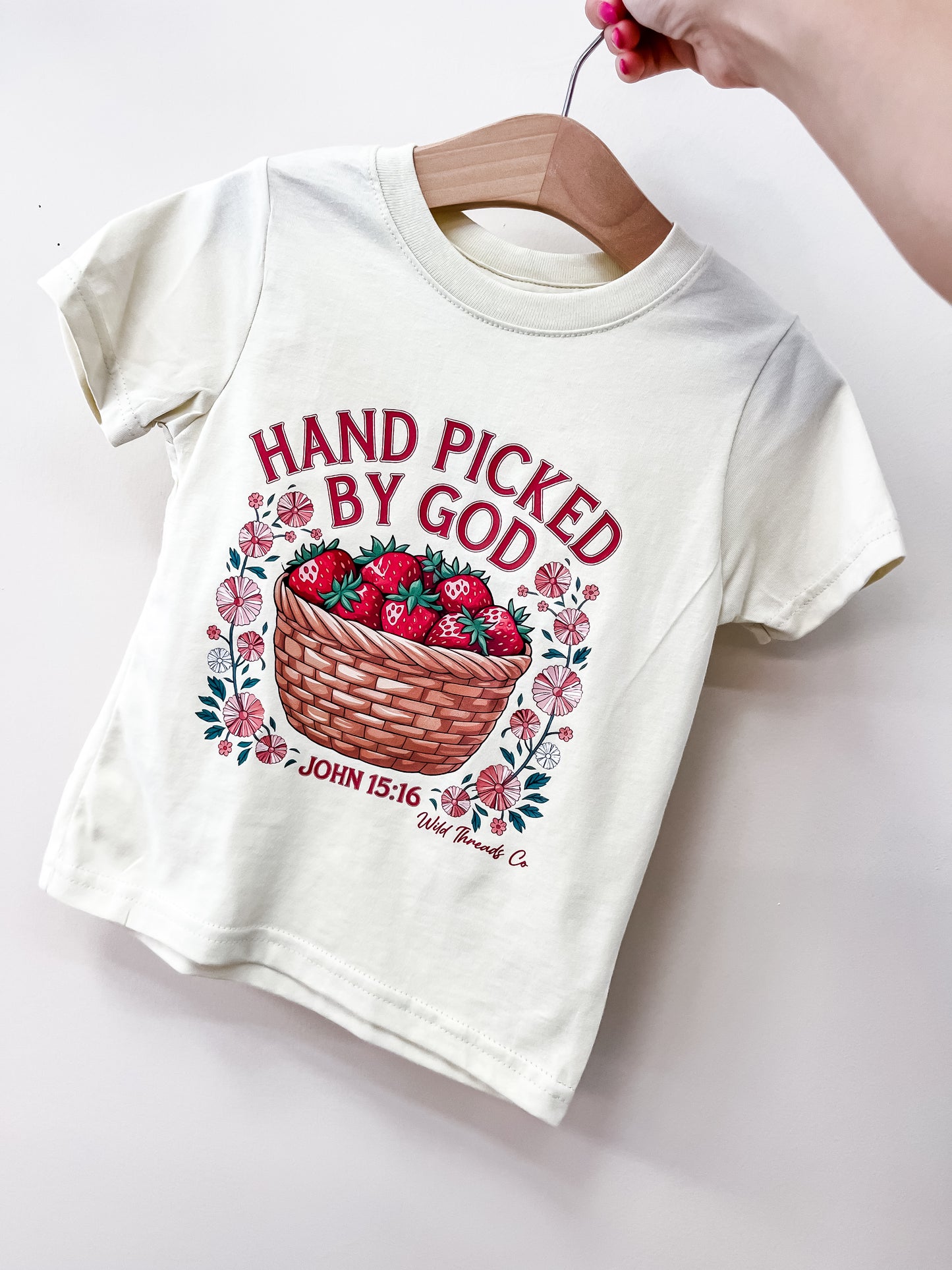 The Hand Picked By God T-Shirt by Wild Threads Co is a white toddler tee on a hanger, featuring a strawberry basket, pink flowers, and red "HAND PICKED BY GOD, JOHN 15:16" text. A hand holds the hanger against a plain background.