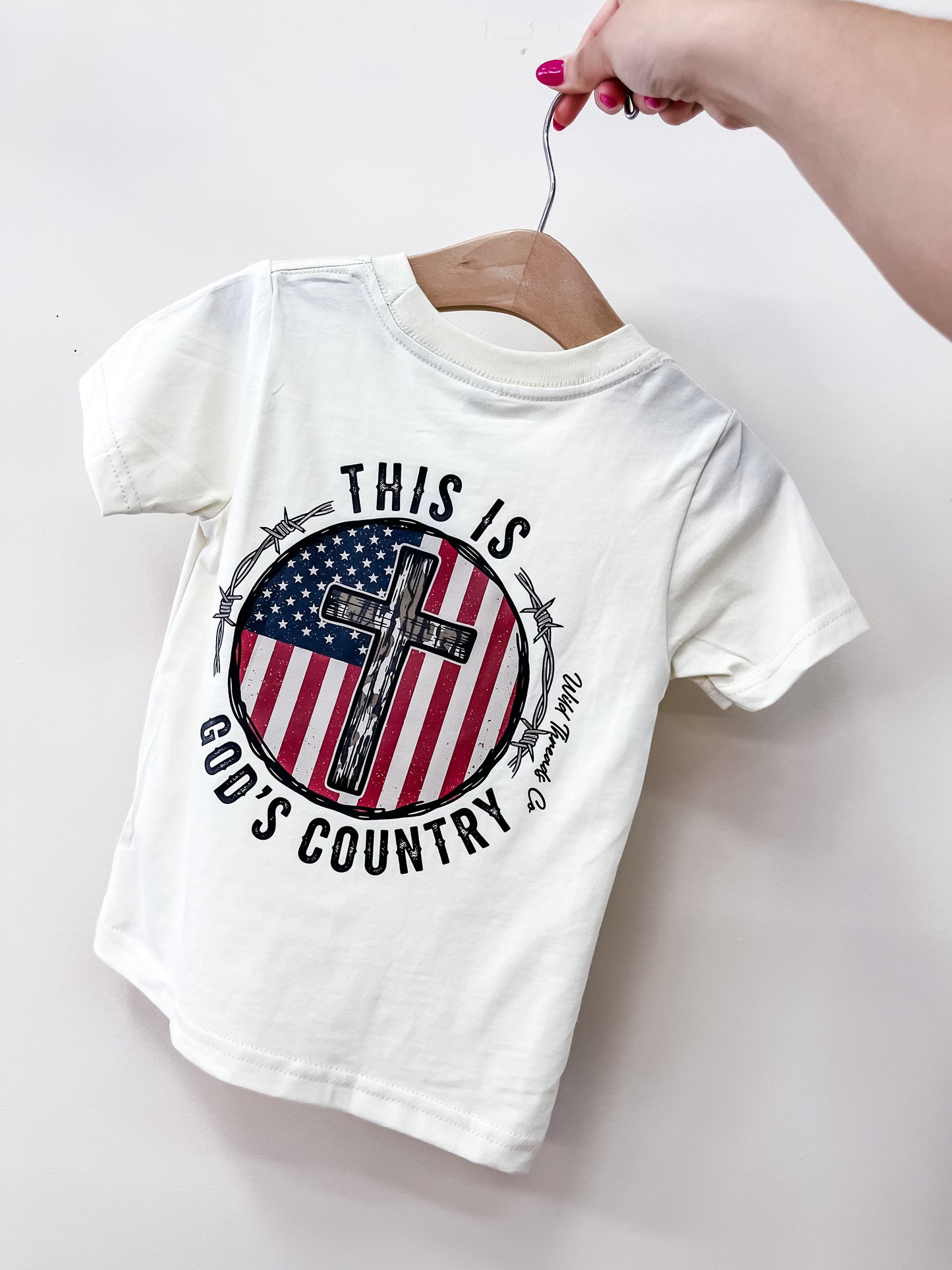A hand with pink nails holds a white God's Country T-Shirt from Wild Threads Co, featuring an American flag in a circle with a cross and barbed wire. The text reads, “THIS IS GOD’S COUNTRY.”.