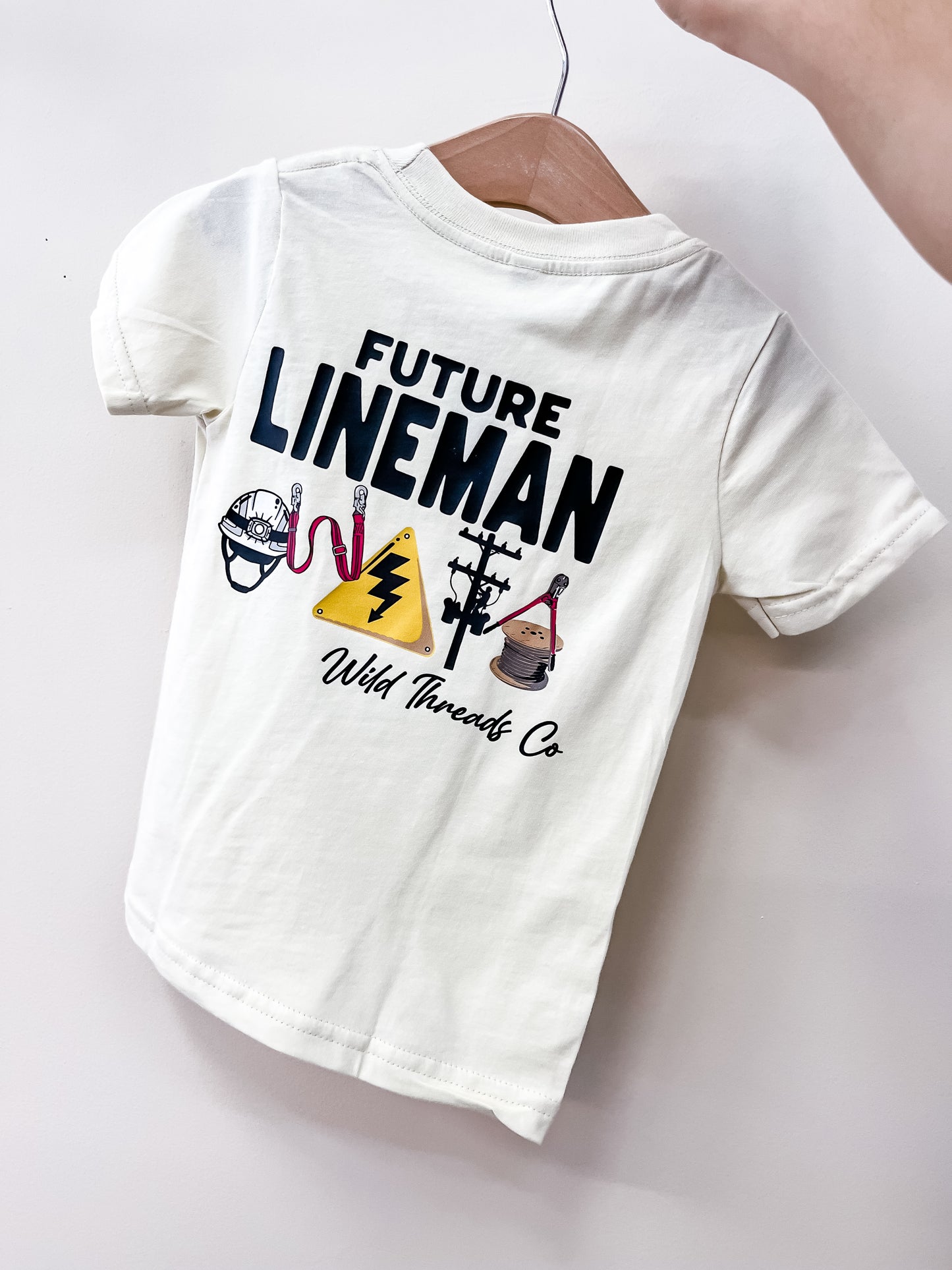 A white toddler Future Lineman T-Shirt by Wild Threads Co hangs on a hanger, featuring FUTURE LINEMAN text with drawings of a hard hat, electrical signs, gloves, bucket truck, and utility pole.