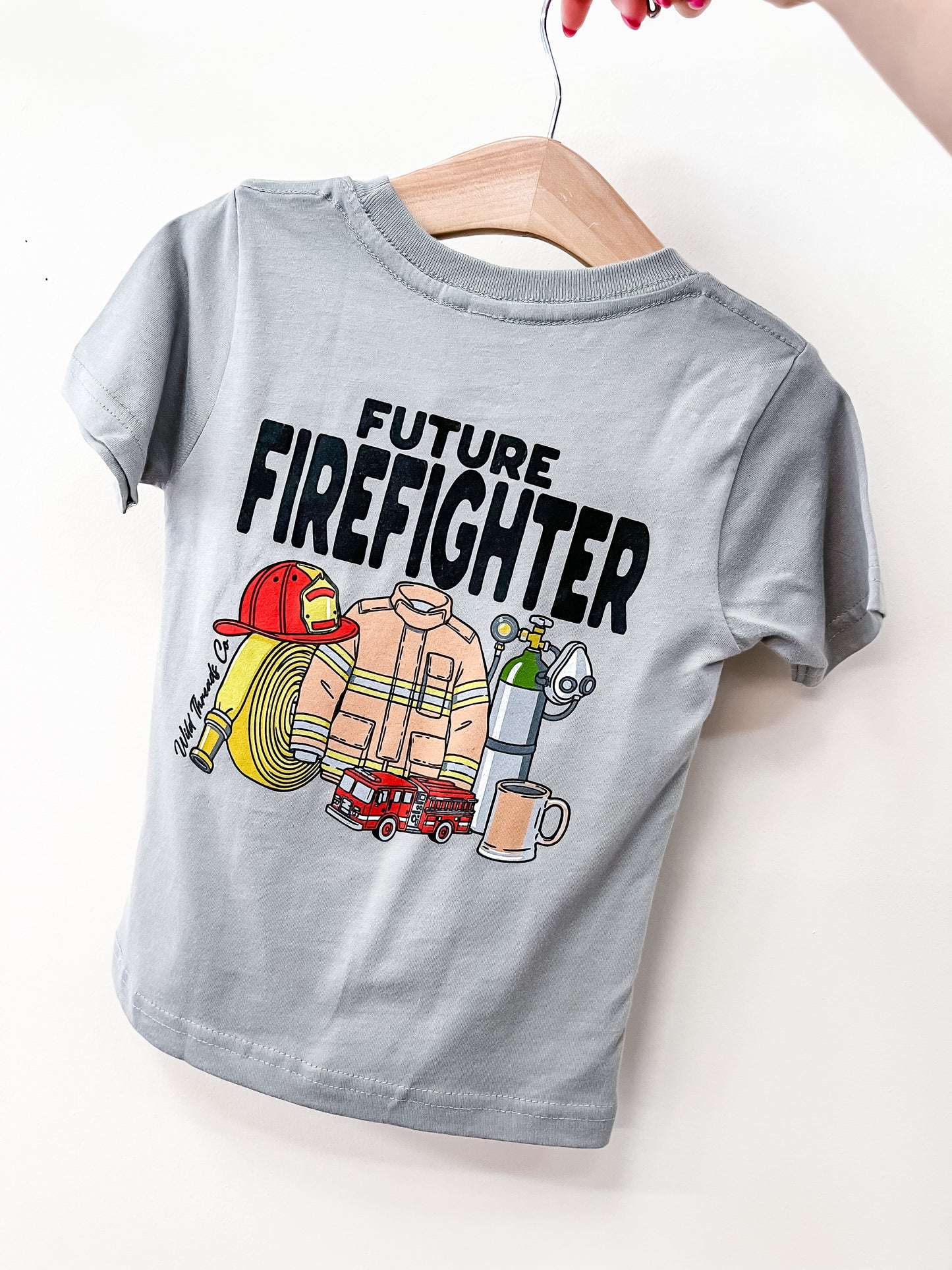 The Wild Threads Co Future Firefighter T-Shirt, shown in gray on a hanger held by a hand, displays "Future Firefighter" with illustrations of a helmet, hose, jacket, oxygen tank, fire truck, and mug.
