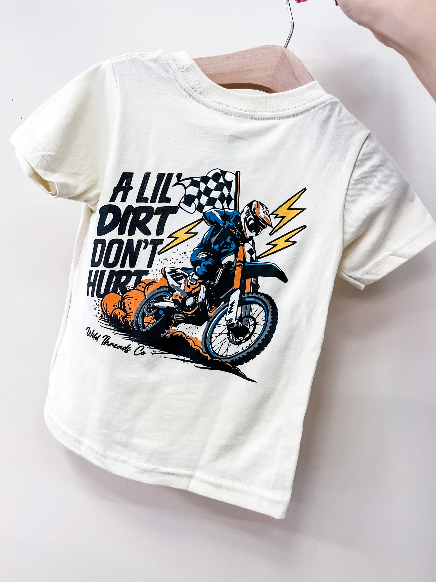 The Wild Threads Co A Lil Dirt T-Shirt is a white kids’ tee featuring a dirt bike rider, lightning bolts, and a checkered flag with “A LIL DIRT DON’T HURT” text. Shown held by hand on a hanger.