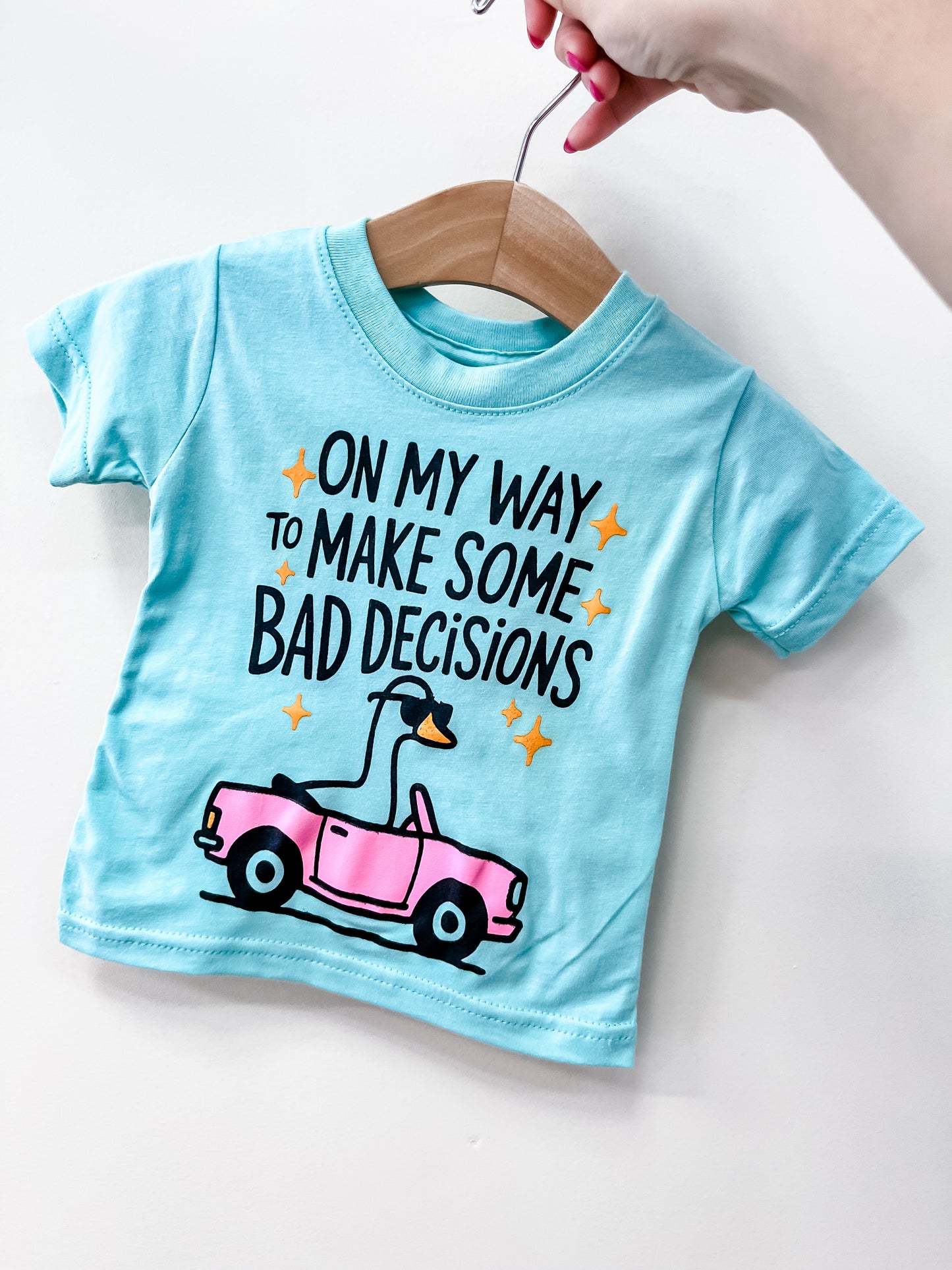 The Bad Decisions t-Shirt by Wild Threads Co is a light blue toddler tee featuring a cartoon goose in a pink convertible and the phrase “On my way to make some bad decisions” printed on the front, shown hanging on a hanger.