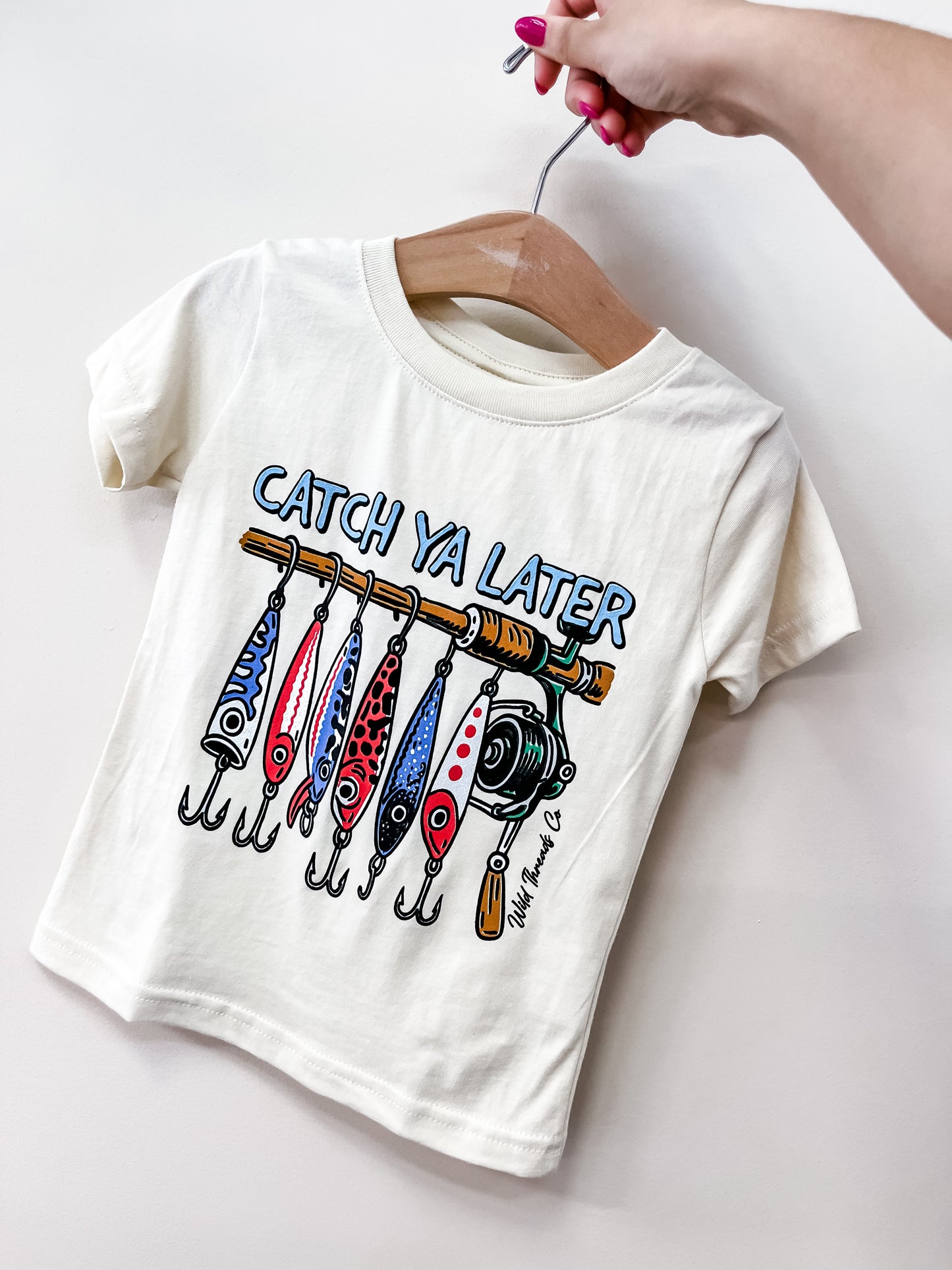 A hand holds a Catch Ya Later T-Shirt by Wild Threads Co, featuring colorful fishing lures and a reel graphic, with “Catch Ya Later” in blue above the design.