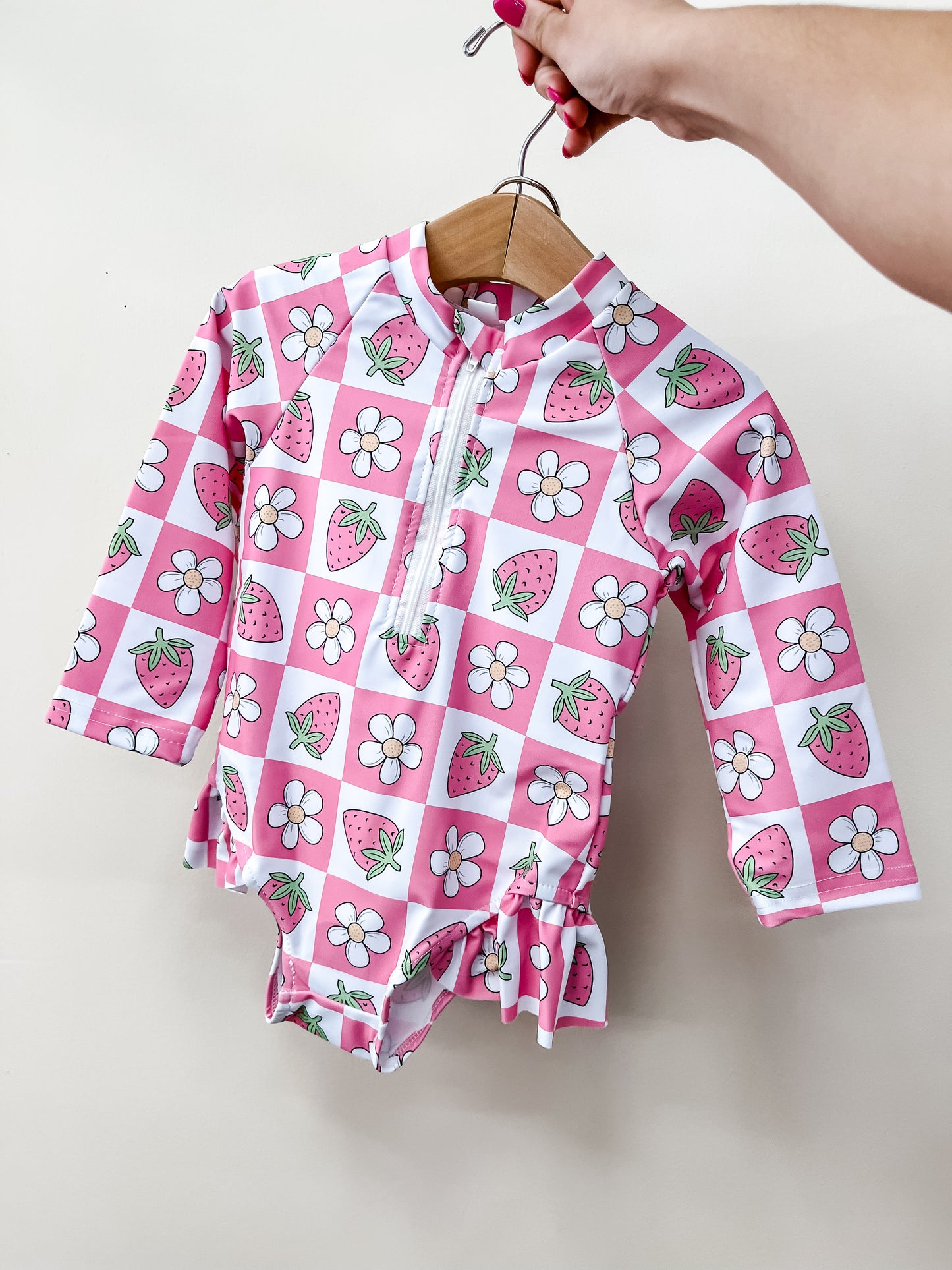 A hand holds the Tiny Tot Bundles Strawberry Check Swimsuit, a pink and white checkered long-sleeve baby swimsuit decorated with strawberries and white flowers, against a plain light background.