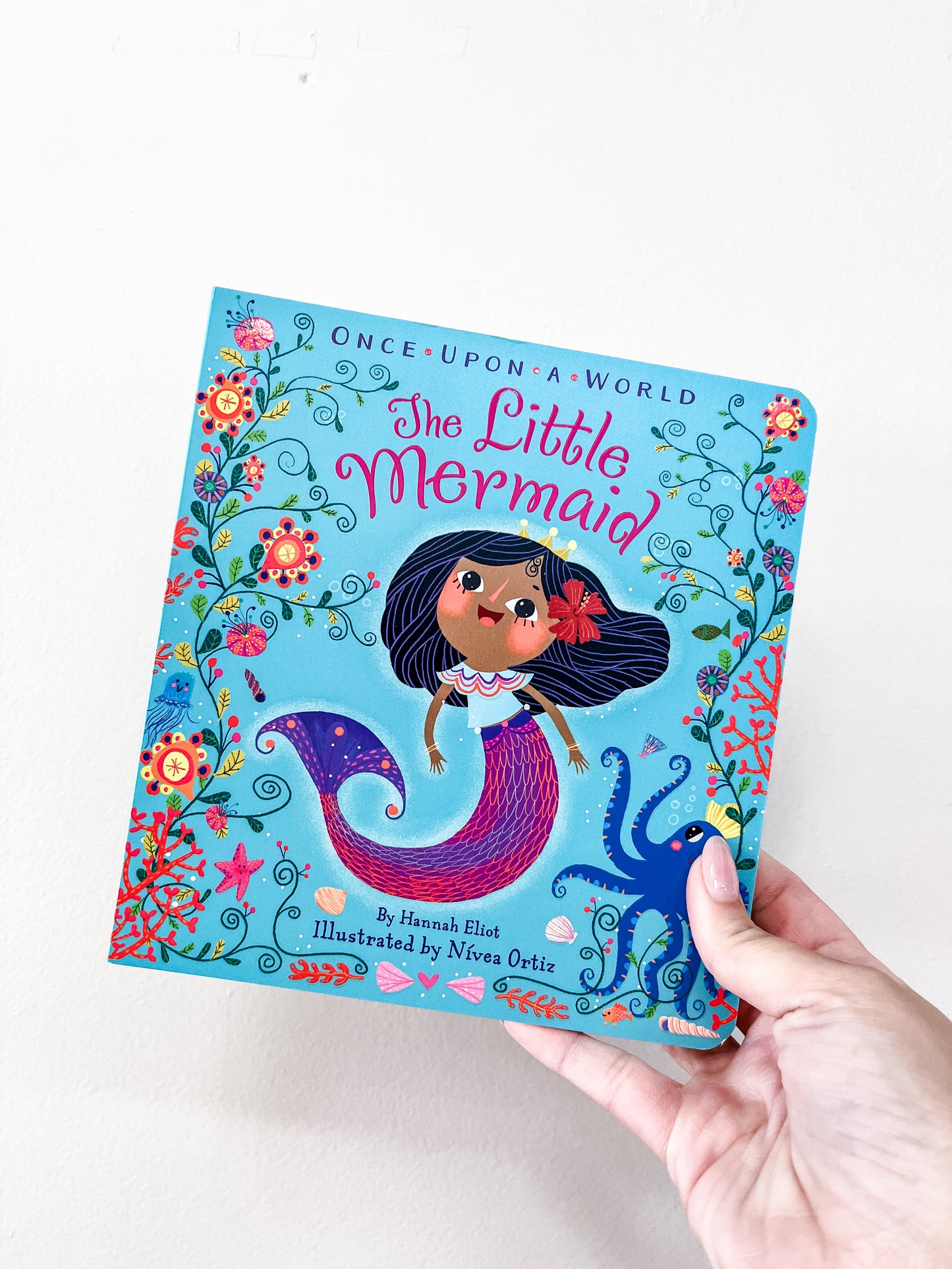 A hand holds the "Little Mermaid Book" by Simon & Schuster, featuring a colorful cover illustration of a dark-haired mermaid surrounded by flowers.