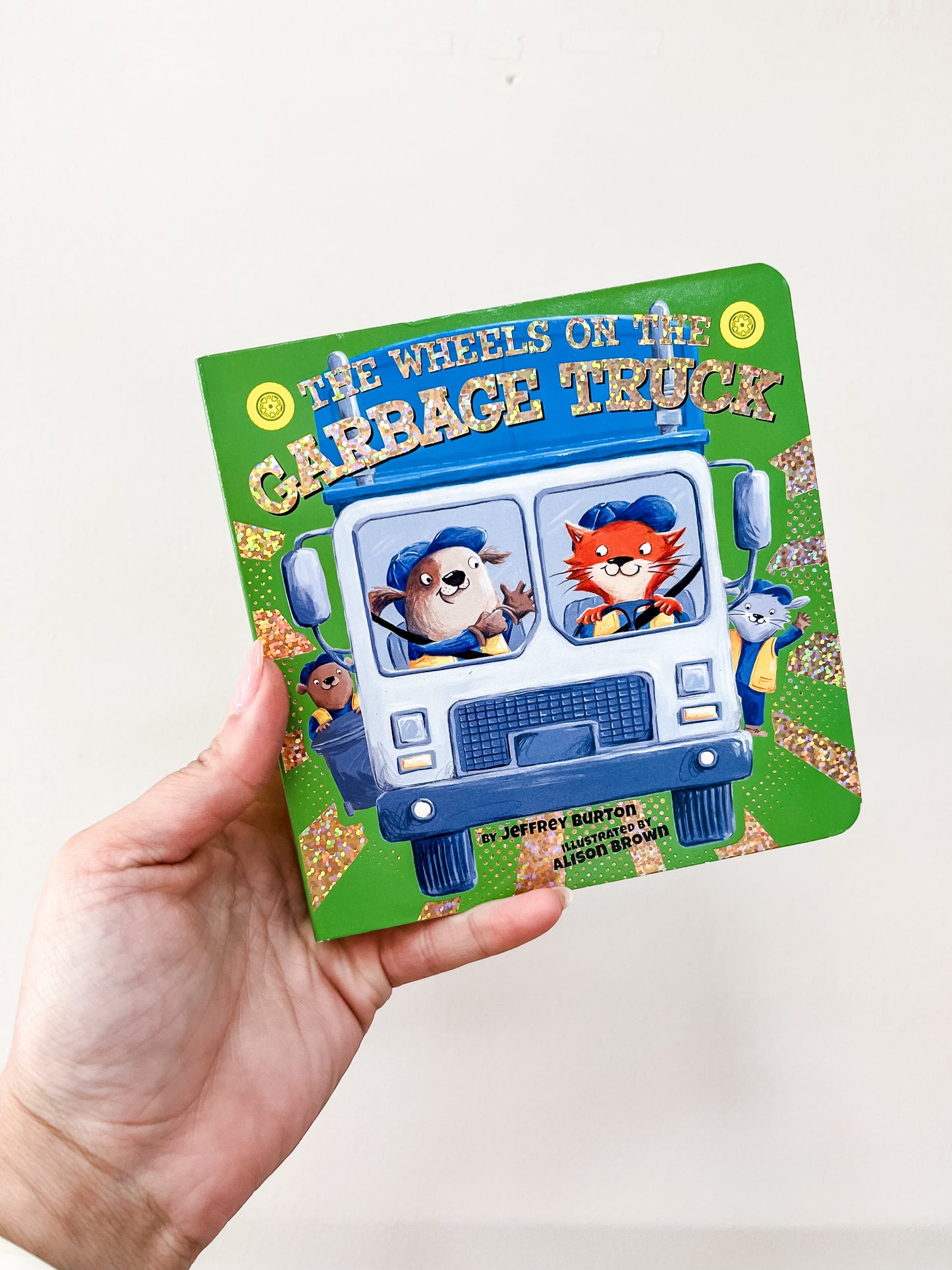 A hand holds the Wheels On the Garbage Truck Book by Simon & Schuster, showing cartoon animals in uniforms driving a blue and white garbage truck on a green cover.