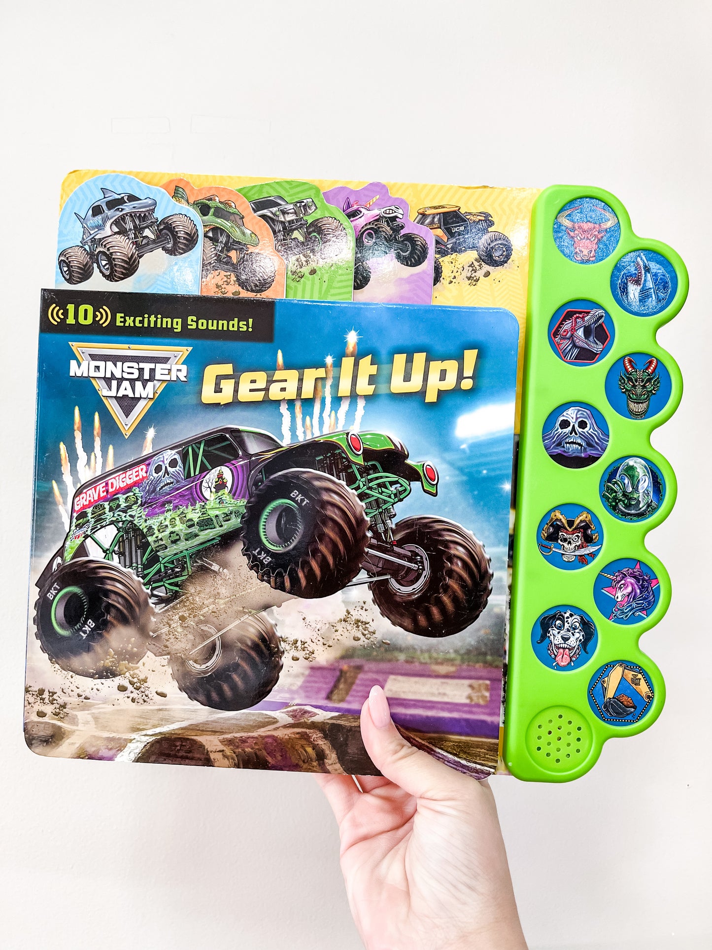A hand holds the Monster Jam: Gear It Up Book by Simon & Schuster, featuring a monster truck on the cover and side buttons with images of monster trucks and related icons.