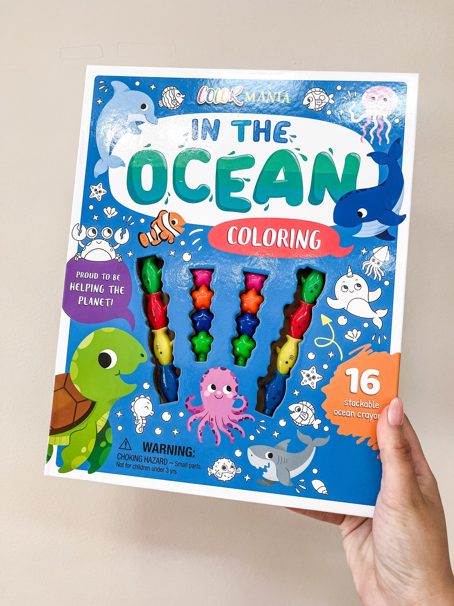 A hand holds a Simon & Schuster In the Ocean Coloring Set Book featuring colorful sea creatures like a turtle, octopus, and fish, with 16 stackable ocean crayons displayed on the front.