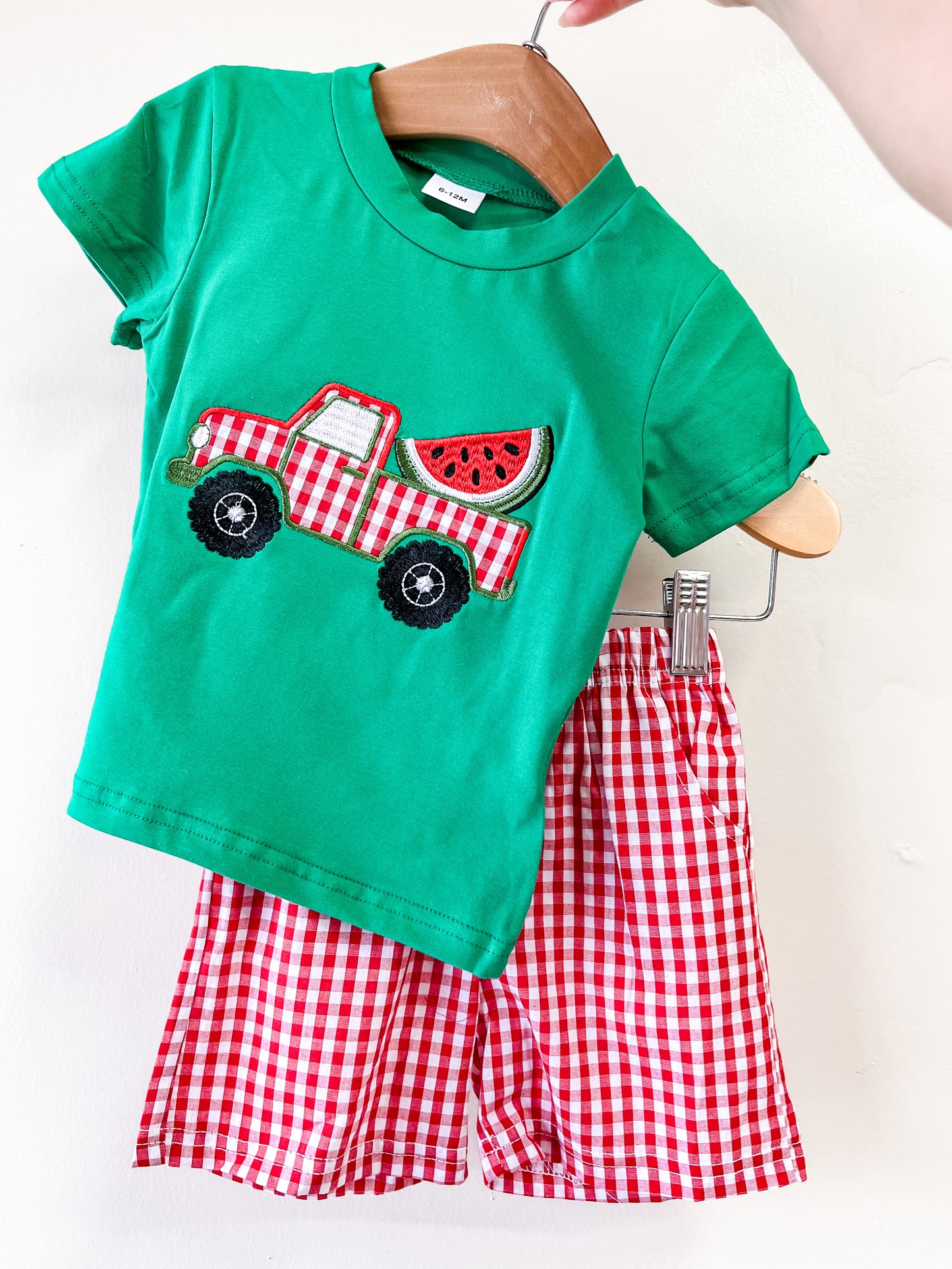 The Watermelon Wagon Set by Tiny Tot Bundles features a green short-sleeve tee with a red plaid truck and watermelon graphic, paired with red and white gingham shorts, displayed on a wooden hanger against a white background.