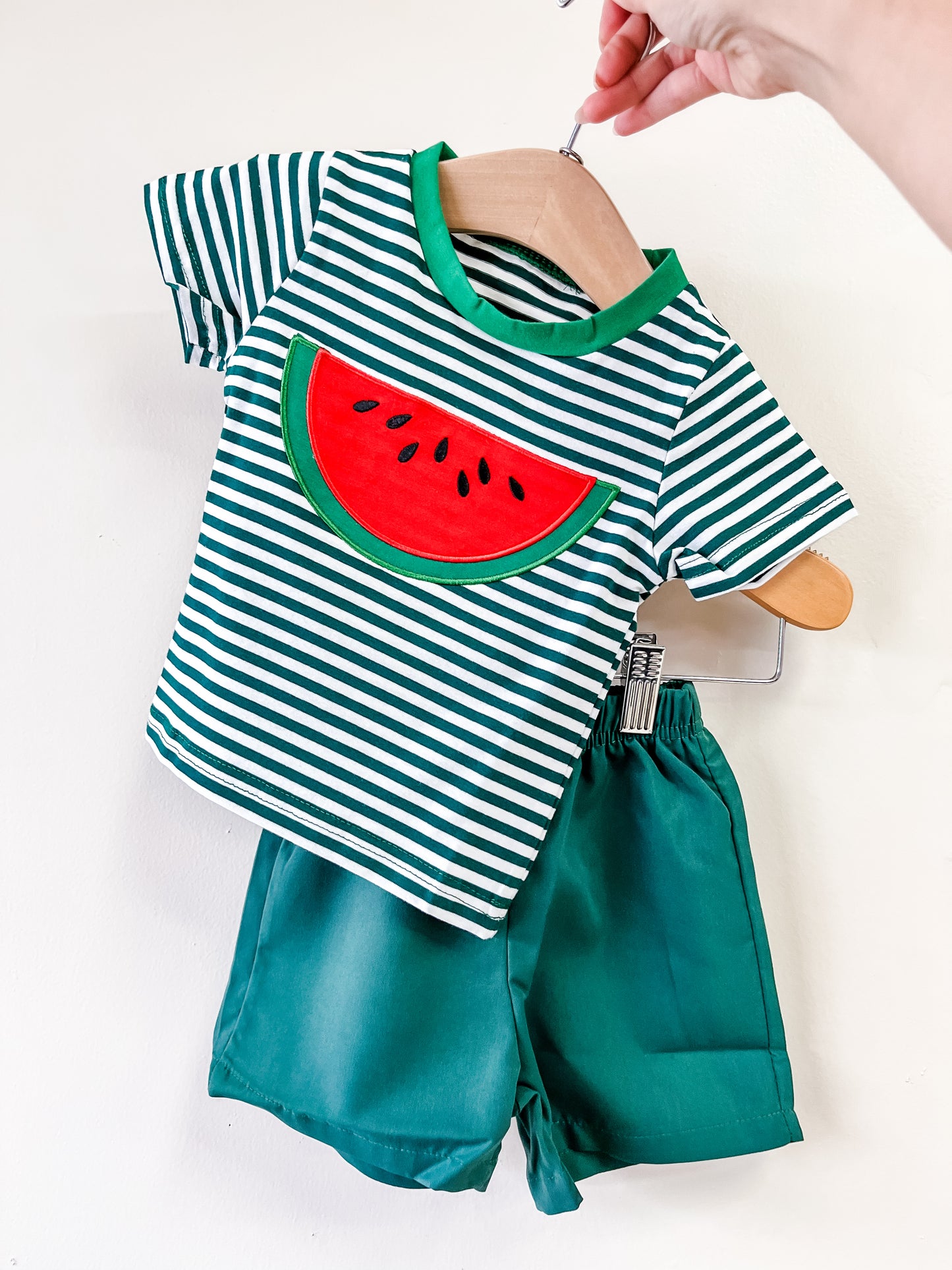 A hand displays the Tiny Tot Bundles Watermelon Adventure Set: a green and white striped t-shirt with a watermelon slice applique, paired with solid green shorts, all shown against a plain white background.