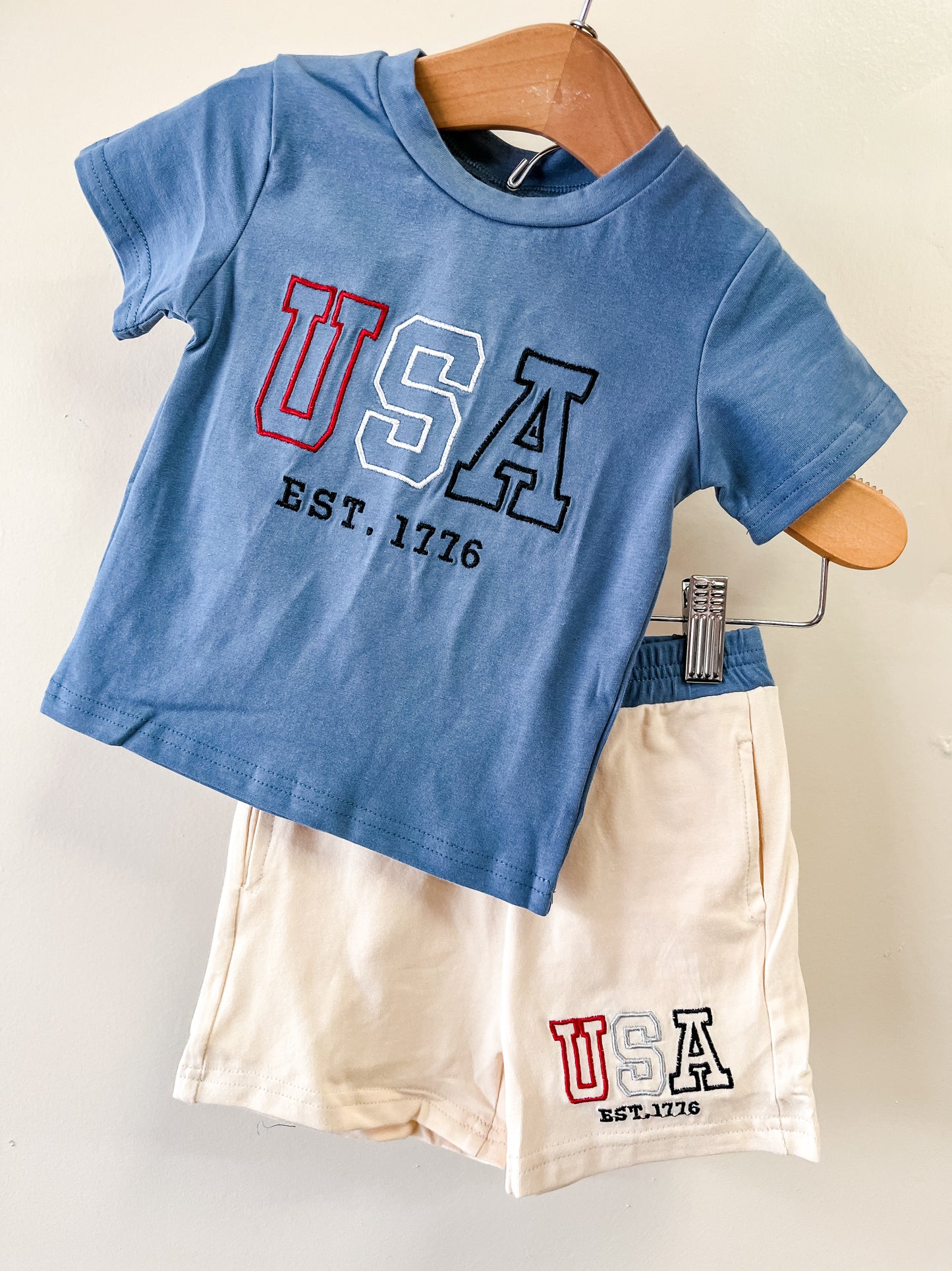 The Little Patriot Set by Tiny Tot Bundles features a blue tee and cream shorts, both with USA EST. 1776 designs, displayed together on a wooden hanger against a light background.