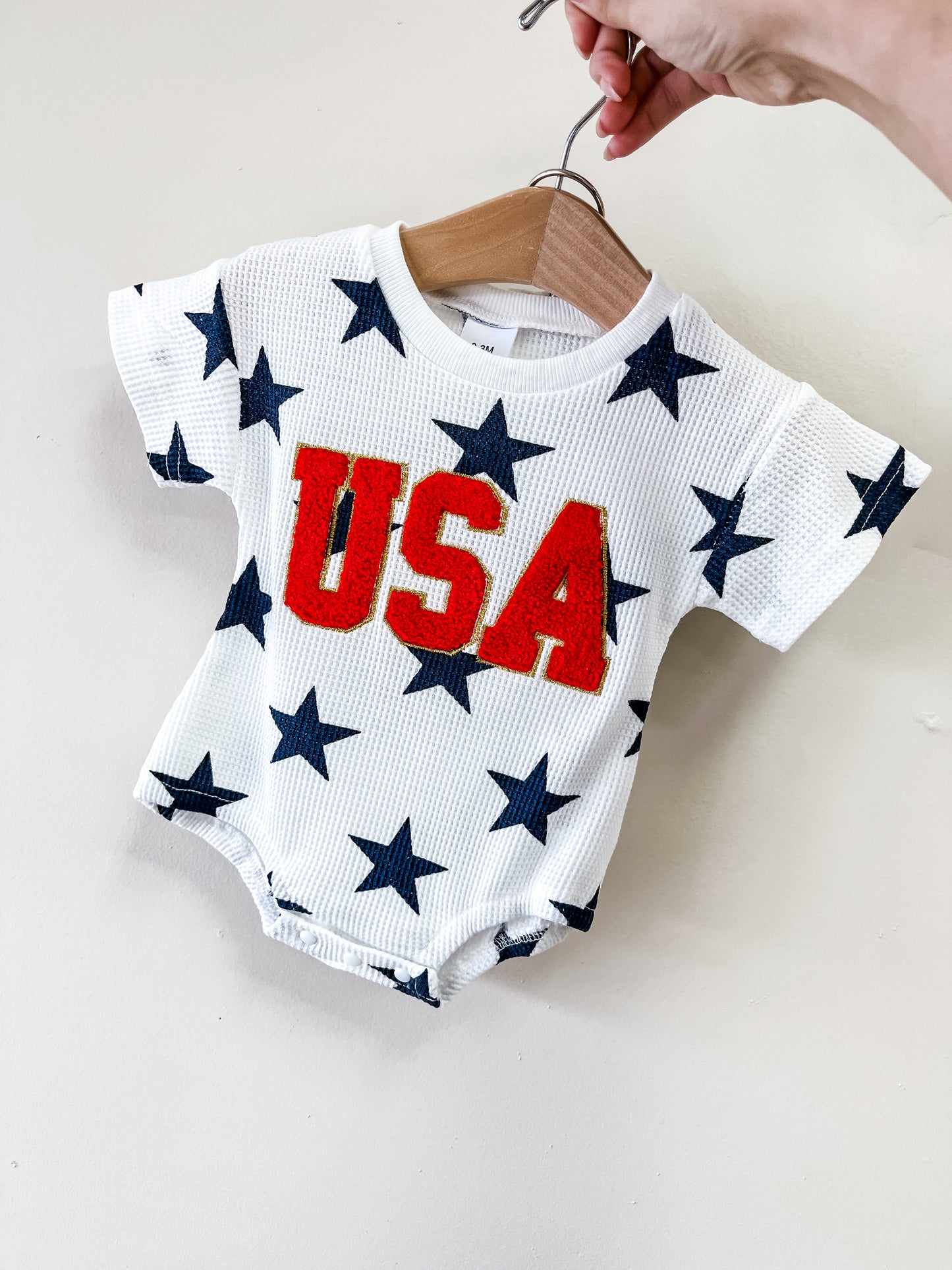 A hand holds the Tiny Tot Bundles Little Firecracker Bubble onesie on a hanger. The white baby outfit features large blue stars and bold red "USA" lettering on the front.