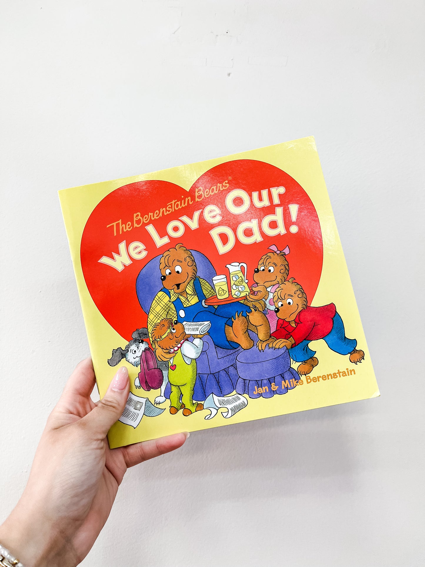 A hand holds the "We Love Our Dad! Book" by Harper Collins, showing the bear family and their father in a blue chair with a big red heart in the background.