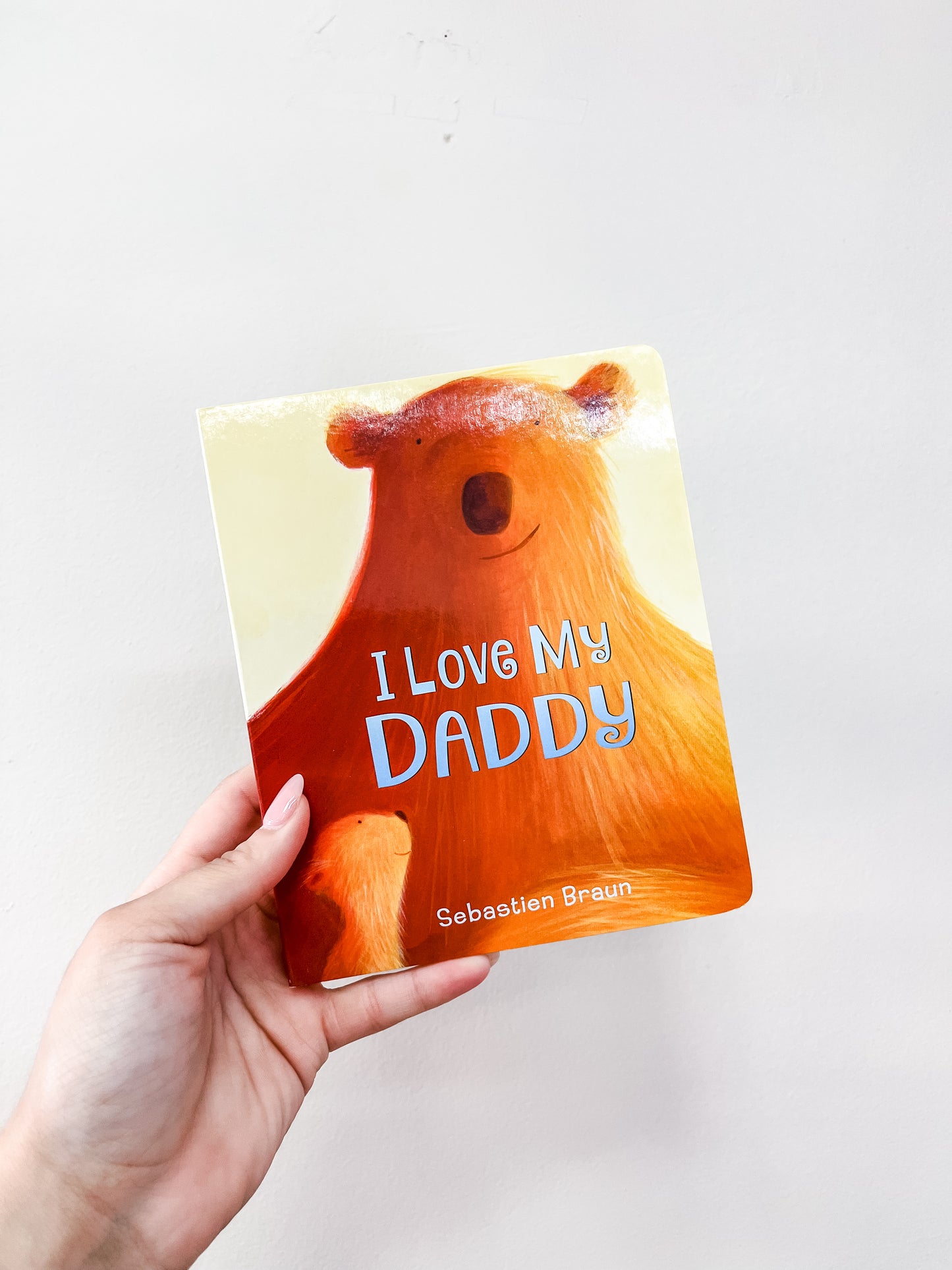 A hand holds the I Love My Daddy Board Book by harper Collins, featuring a warm orange-toned illustration of a bear on the cover.