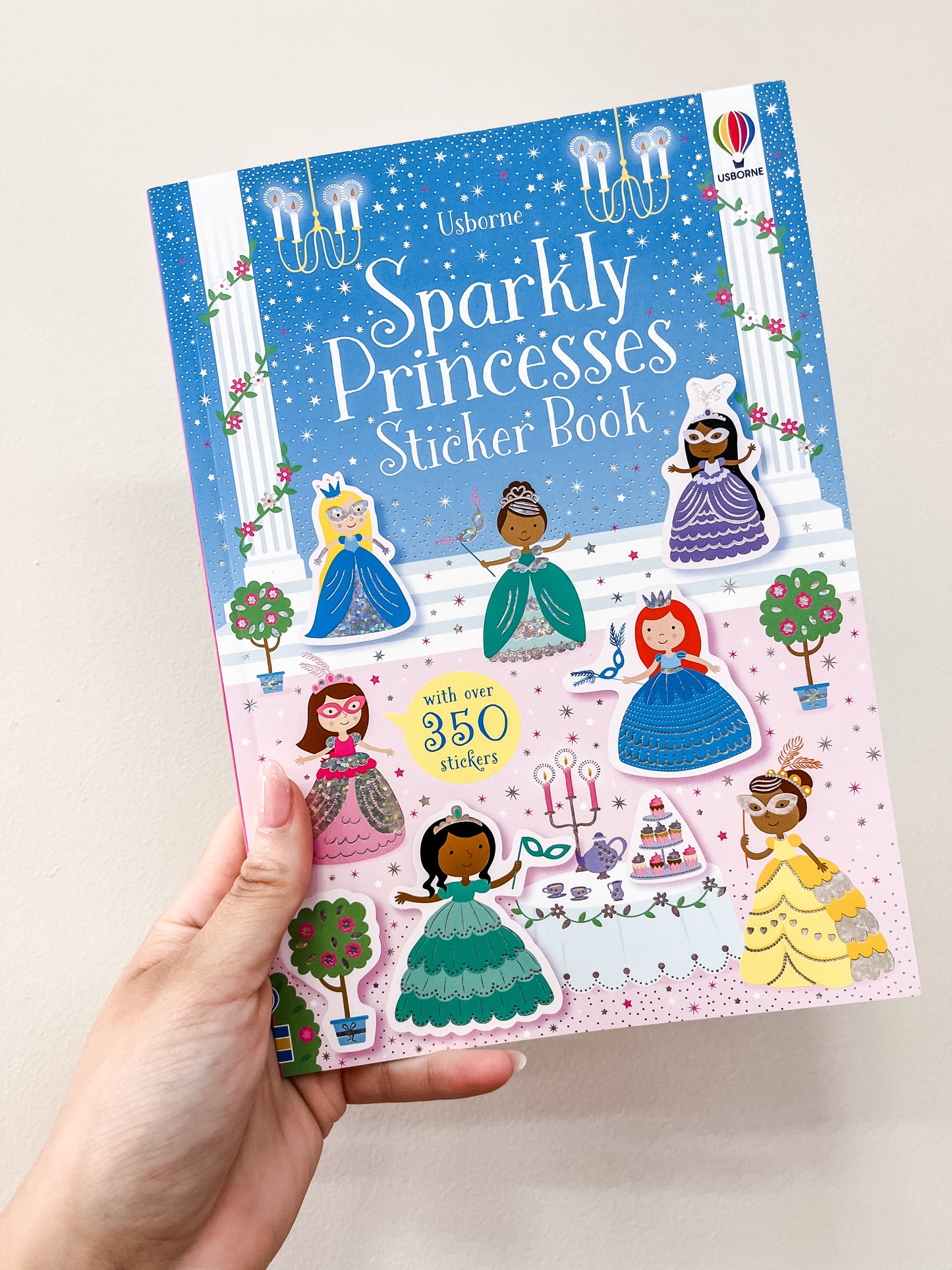A hand holds the Sparkly Princesses Sticker Book by Harper Collins, featuring illustrated princesses, chandeliers, a garden scene, and over 350 stickers on the colorful cover.