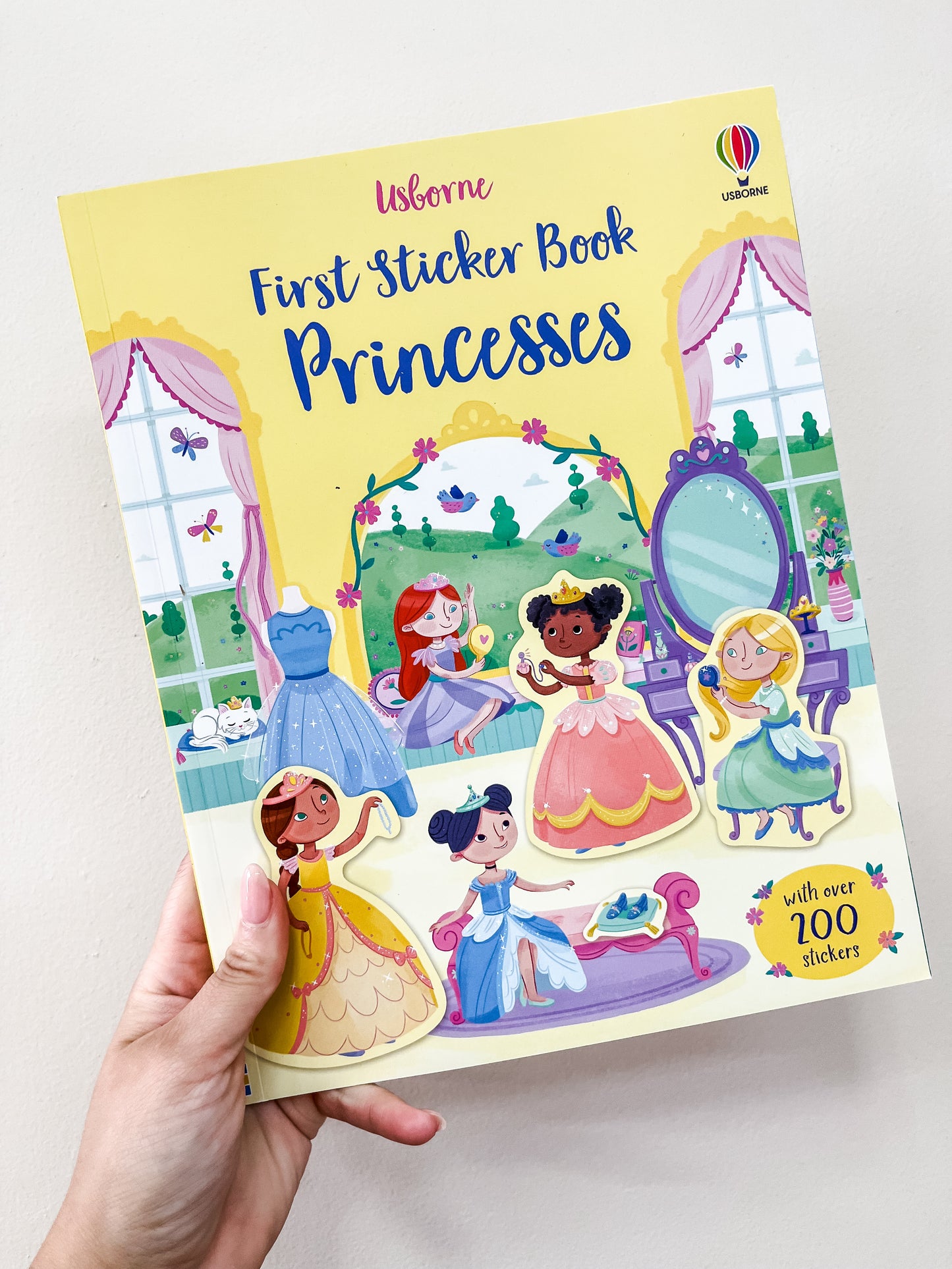 A hand holds the Harper Collins First Sticker Book Princesses, a colorful book showing illustrated princesses, a mirror, and a dressing room; its cover highlights "with over 200 stickers.
