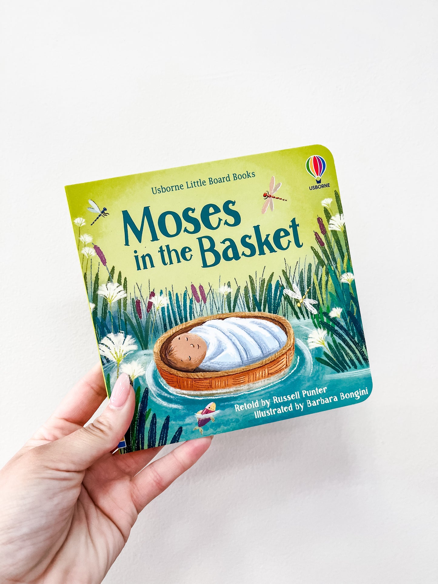 A hand holds the "Moses in the Basket Board Book" by Harper Collins, showing baby Moses wrapped in a blanket, lying in a basket on water, surrounded by grass and flowers.