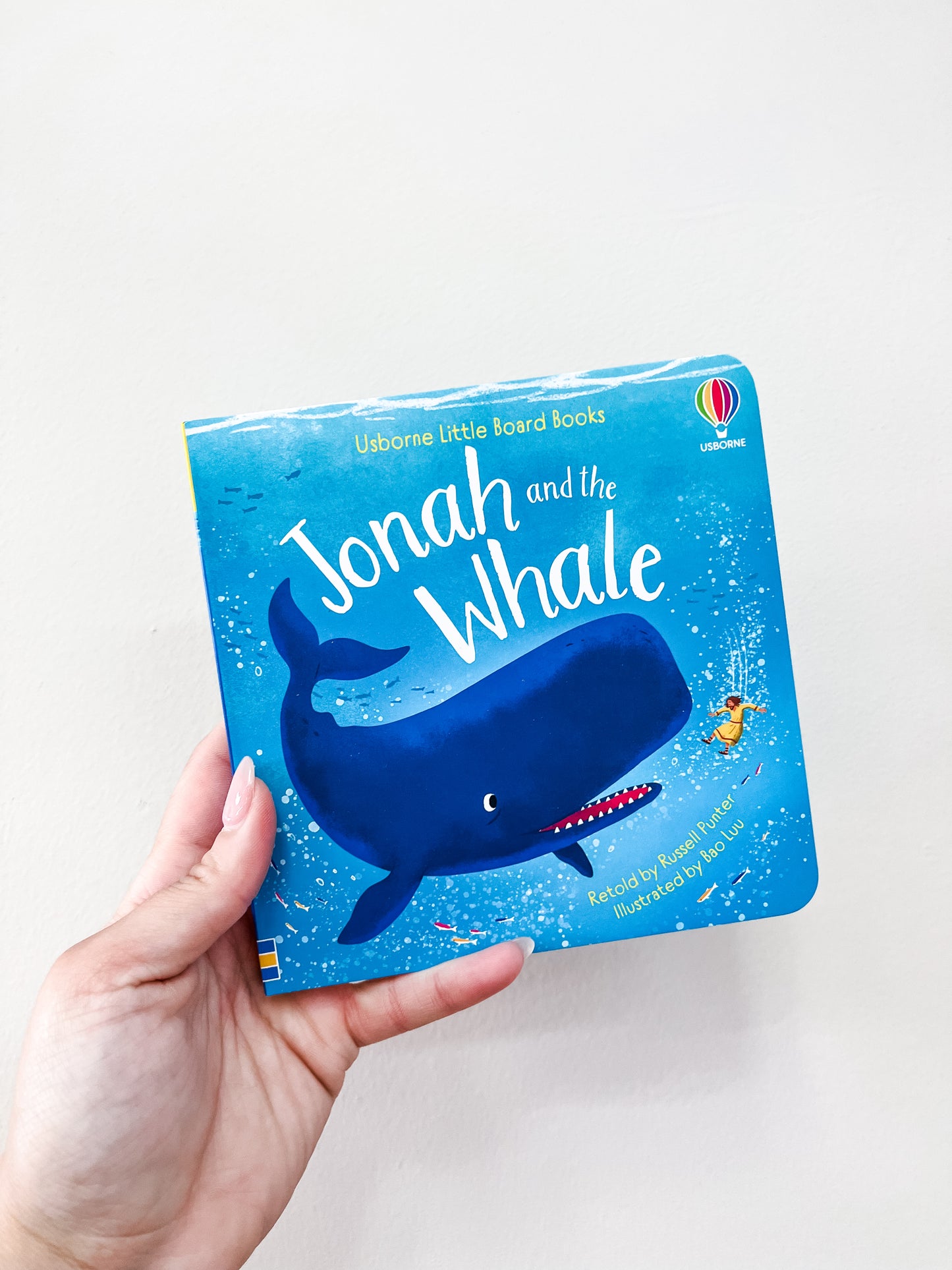 A hand holds the "Jonah and the Whale Board Book" by Harper Collins, showing a blue whale and an orange fish on a blue background with colorful confetti-like dots.