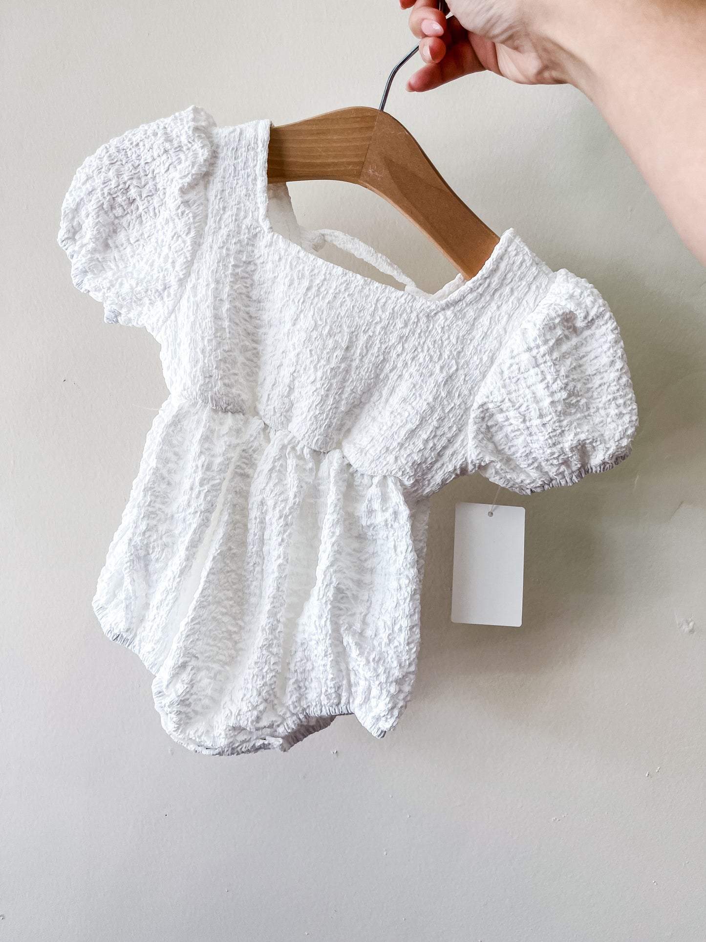 A hand holds a wooden hanger with the Sweet Peony Romper by Tiny Tot Bundles, a white textured baby outfit featuring short puff sleeves, a gathered waist, and a blank white tag attached.