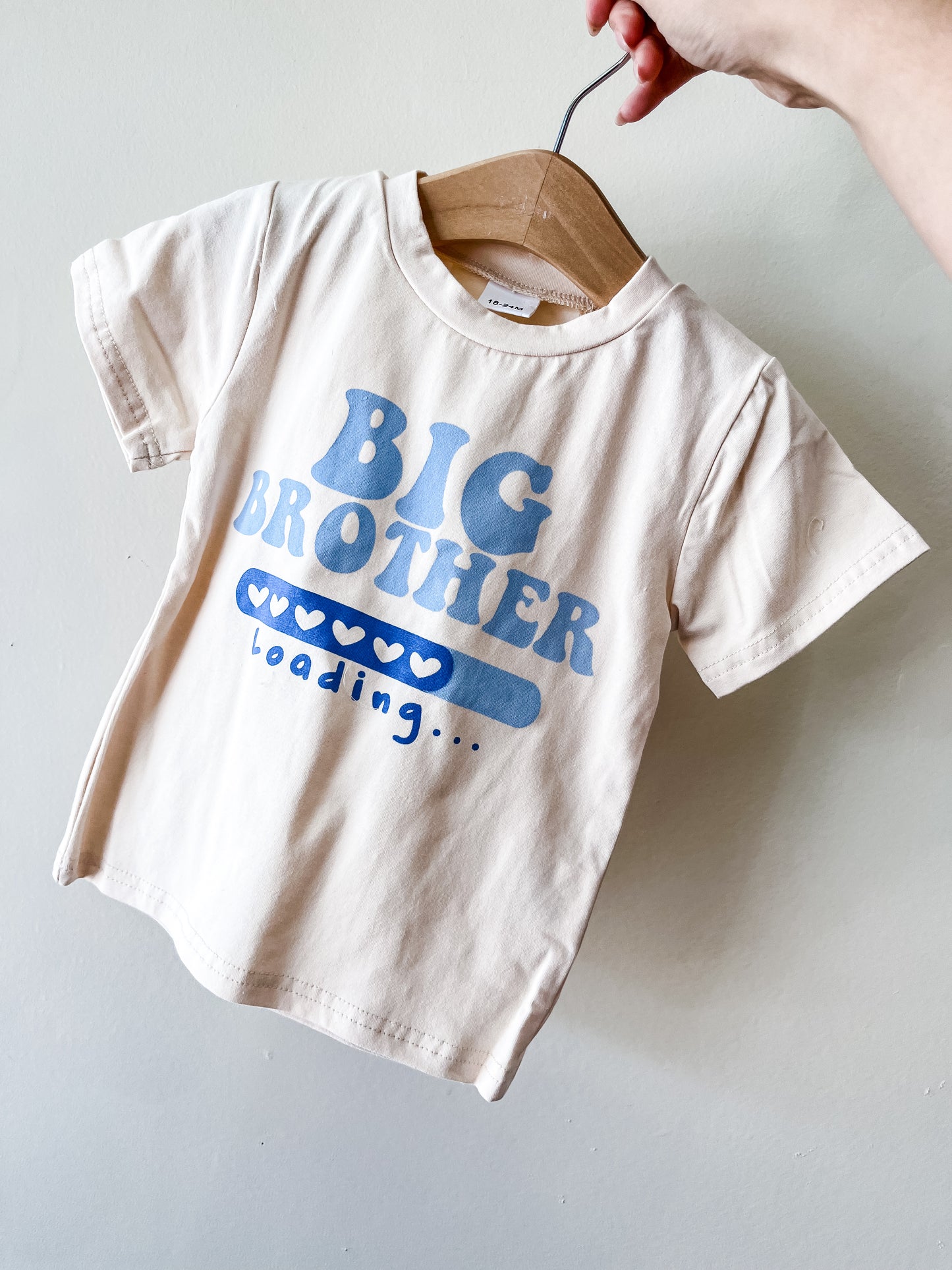 A hand holds a hanger with Tiny Tot Bundles' Big Brother Loading Tee, a light-colored child's T-shirt featuring "BIG BROTHER loading..." in blue and a heart-shaped loading bar under the text.