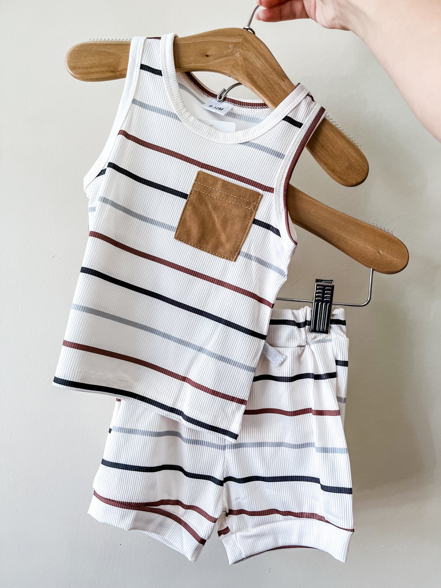 A hand holds a hanger with the Neutral Stripe Tank Set by Tiny Tot Bundles—white with horizontal black, brown, and rust stripes. The sleeveless top features a brown chest pocket and comes with matching shorts.