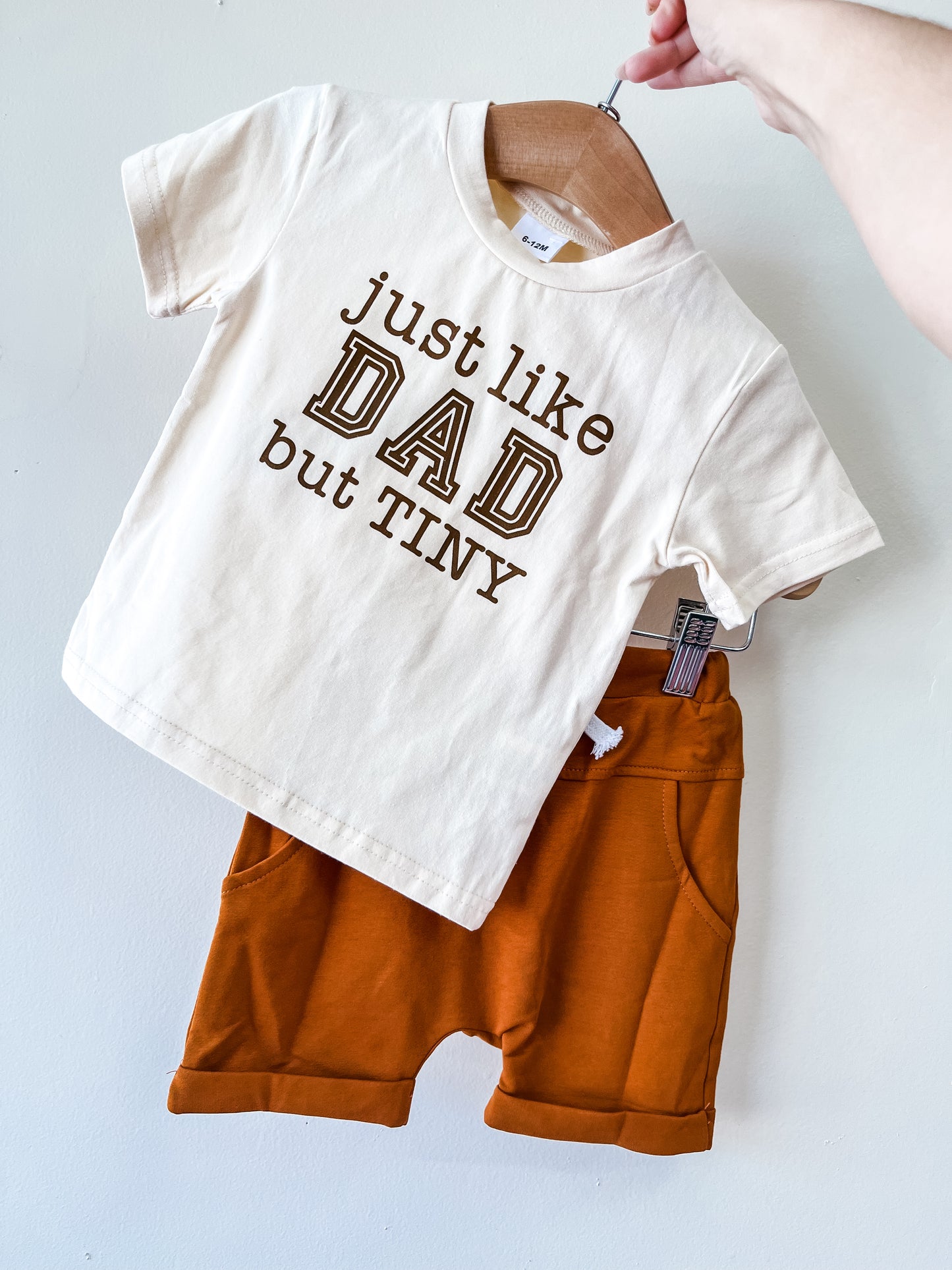 The Just Like Dad Short Set by Tiny Tot Bundles features a cream toddler t-shirt with "just like DAD but TINY" in brown letters and orange shorts, hanging on a wooden hanger against a white background.