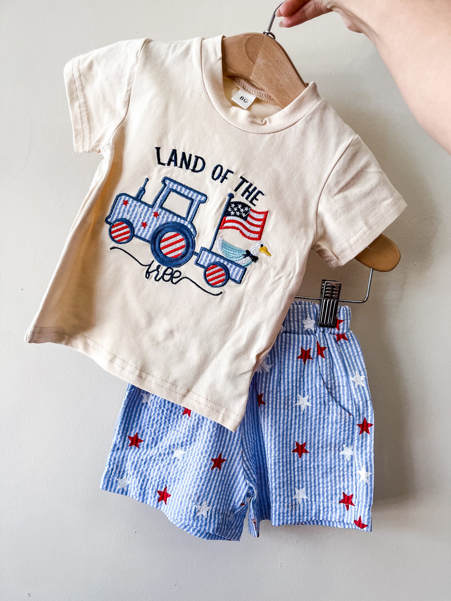 A hand displays the Tiny Tot Bundles "Land Of The Free Set"—a cream tee with a train, American flag, and "Land of the Free" text, paired with blue-and-white striped shorts featuring red and white stars.