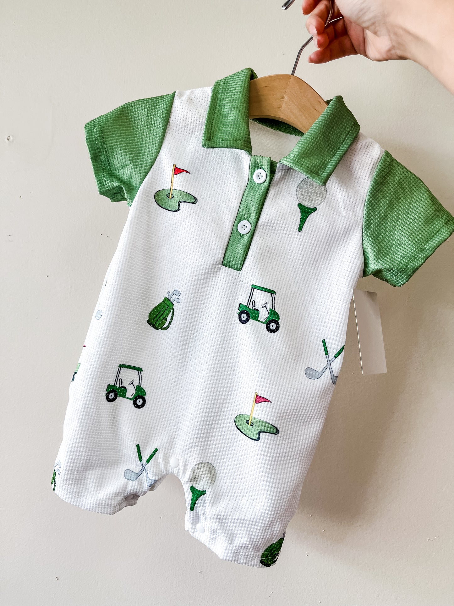 The Tiny Tot Bundles Golf Cart Adventures Romper features green sleeves and collar, with golf carts, flags, tees, clubs, and bags on a white background. Displayed on a wooden hanger held by hand against a light wall.
