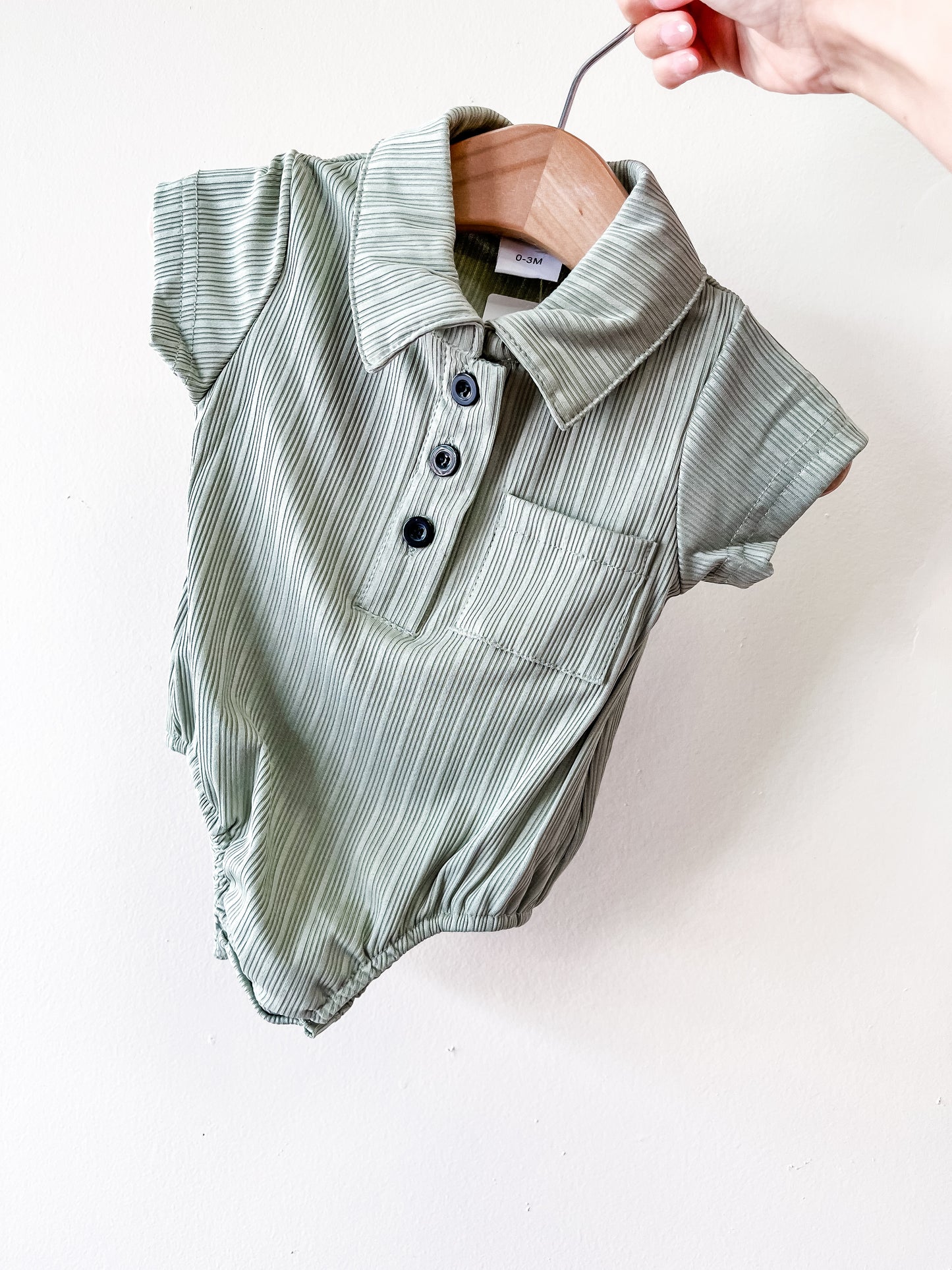 A hand holds the Tiny Tot Bundles Dusty Sage Polo Bubble—a green, short-sleeved baby romper with a collar, front pocket, and three black buttons—on a wooden hanger against a plain white background.