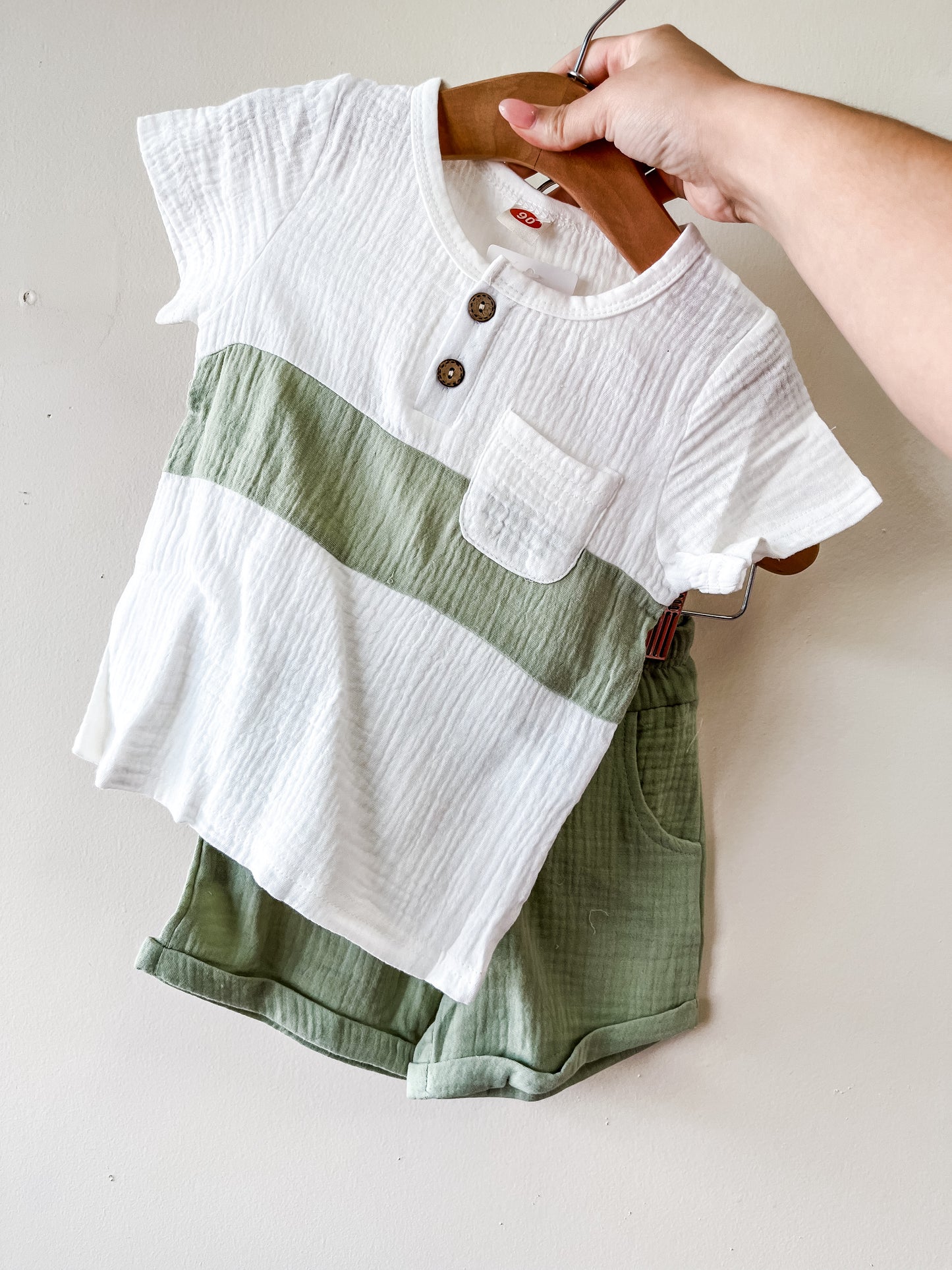 A hand displays the Tiny Tot Bundles Linen White & Green Set, featuring a short-sleeved color-blocked shirt with buttons and a pocket, paired with light green shorts, all on a wooden hanger.