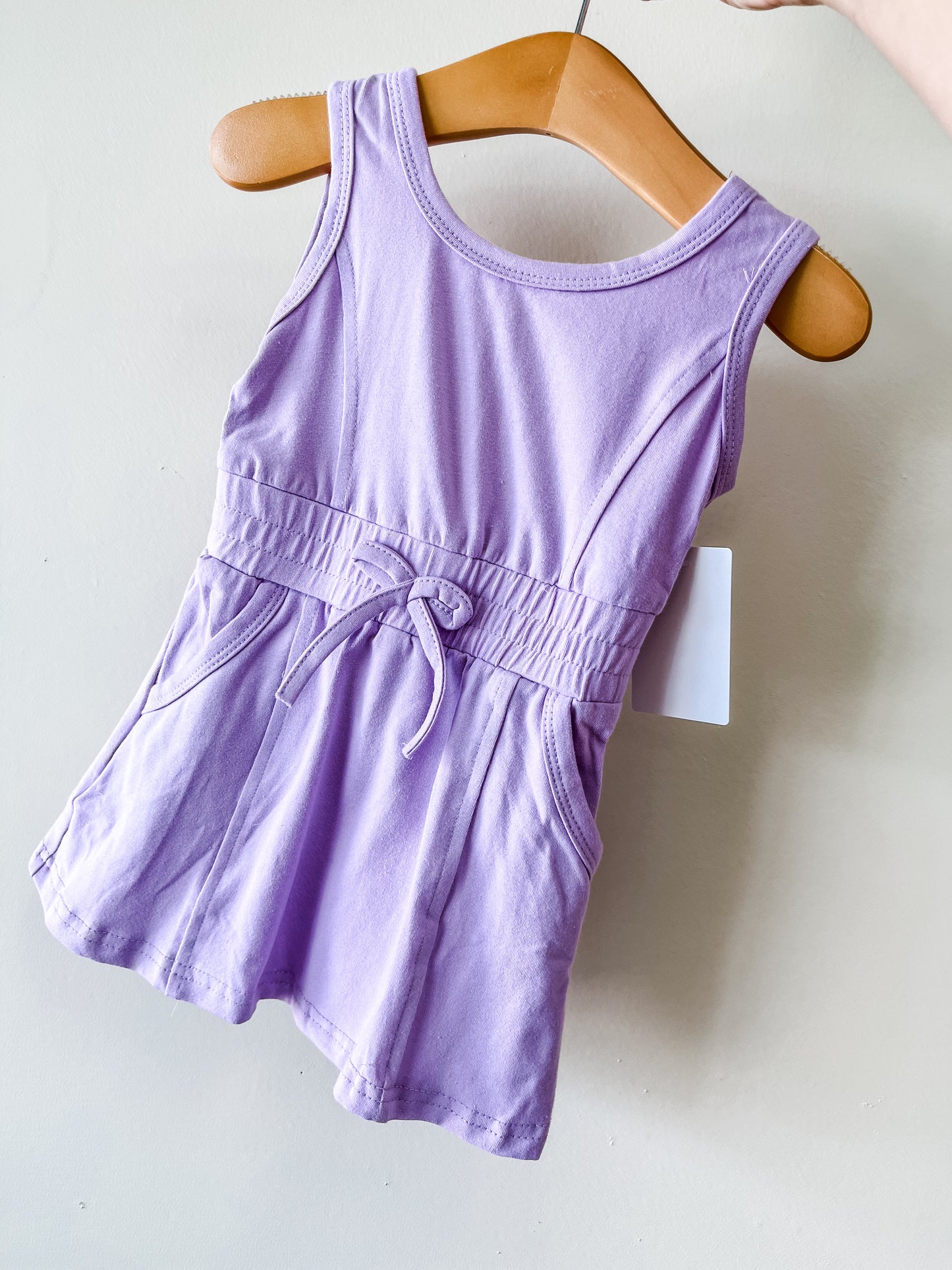 The Tiny Tot Bundles Purple Sporty Dress is a sleeveless toddler dress in light purple with a gathered waist, front bow tie, and two pockets, shown on a wooden hanger against a light background.