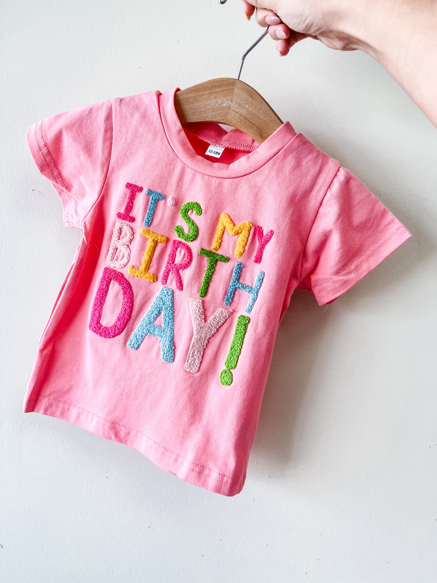 A hand holds a wooden hanger with Tiny Tot Bundles' It's My Birthday Tee—a pink toddler T-shirt featuring colorful, textured "ITS MY BIRTHDAY!" letters—against a white background.