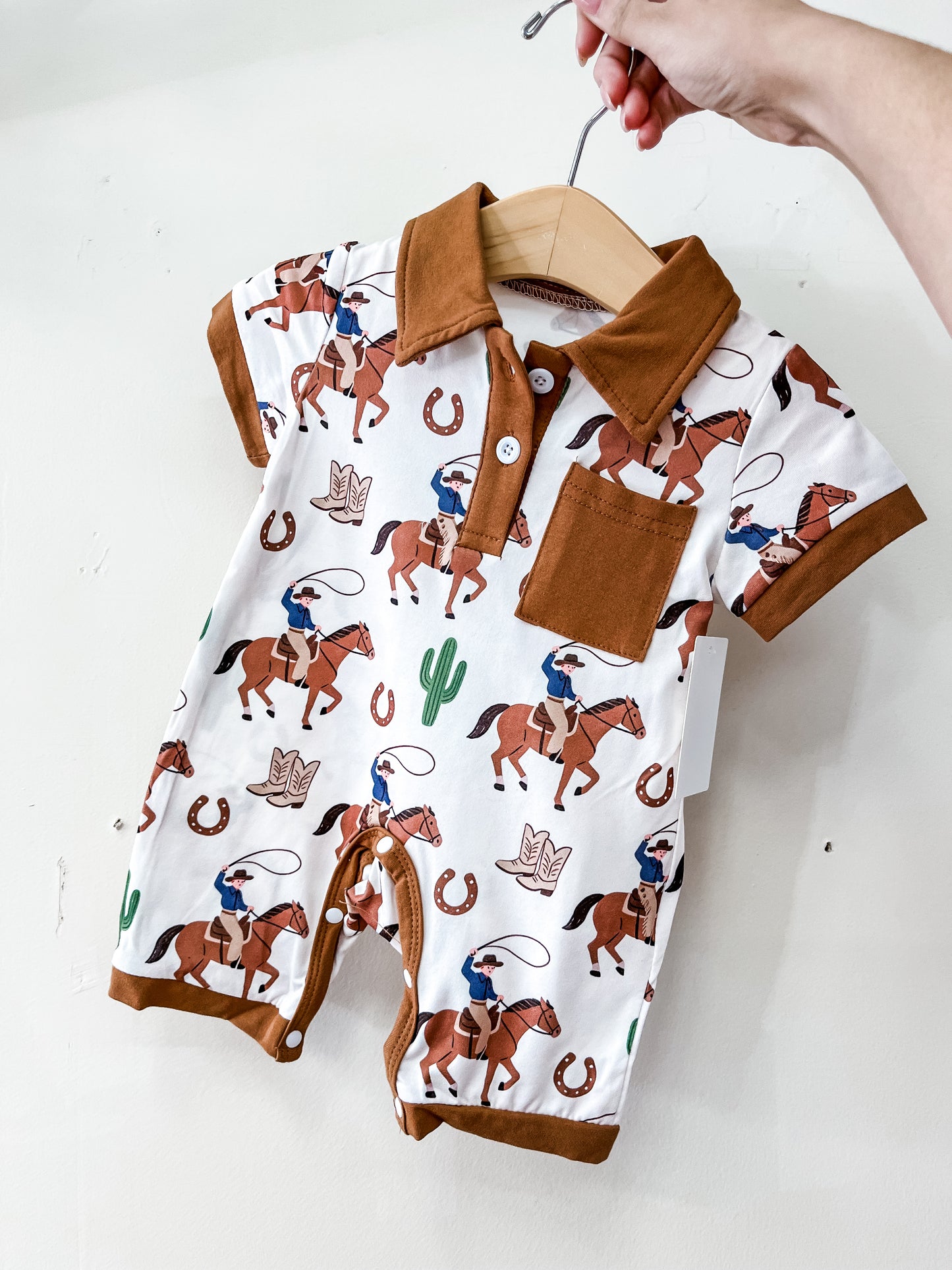 A hand holds up the Tiny Tot Bundles Brown Cowboy Romper, a short-sleeve outfit with brown trim, collar, and front pocket, featuring a fun print of cowboys on horses, cacti, horseshoes, and cowboy hats.