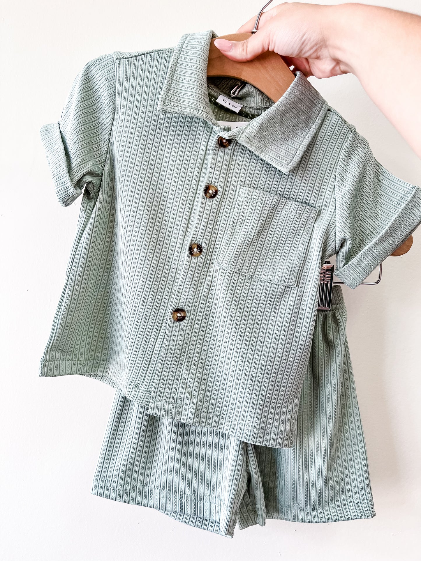 A hand holds up the Tiny Tot Bundles Sage Breeze Set—a ribbed, light green short-sleeve button-up shirt with a front pocket and matching shorts—both hanging on a wooden hanger against a white background.