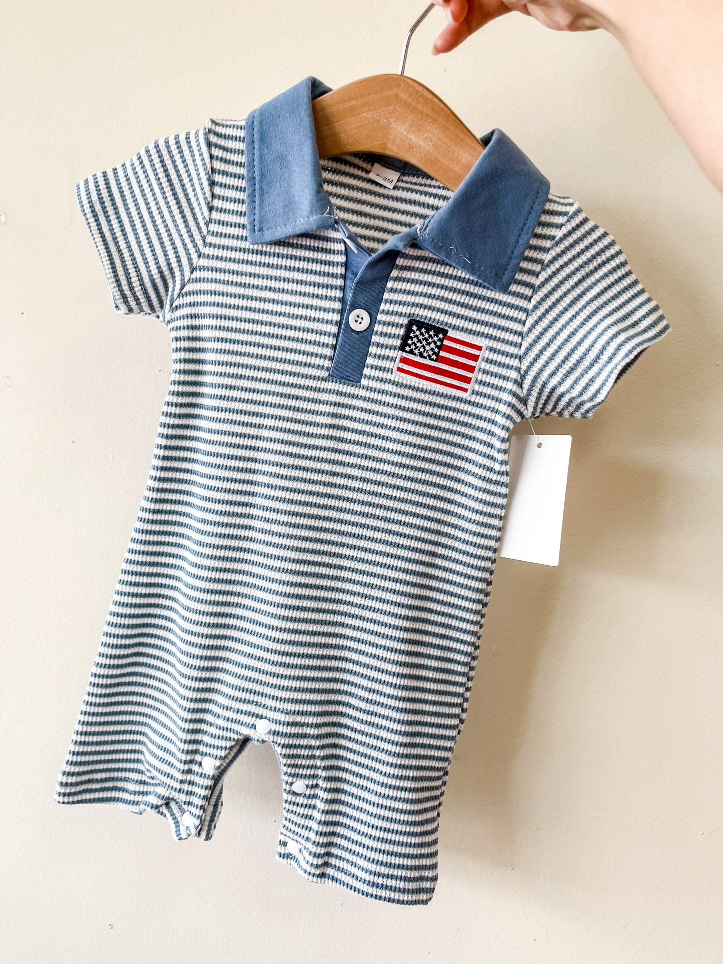 A hand holds up the Tiny Tot Bundles All American Stripe Romper, featuring blue and white stripes, short sleeves, a blue collar, buttons, an embroidered American flag patch on the chest, and a blank white tag on the sleeve.