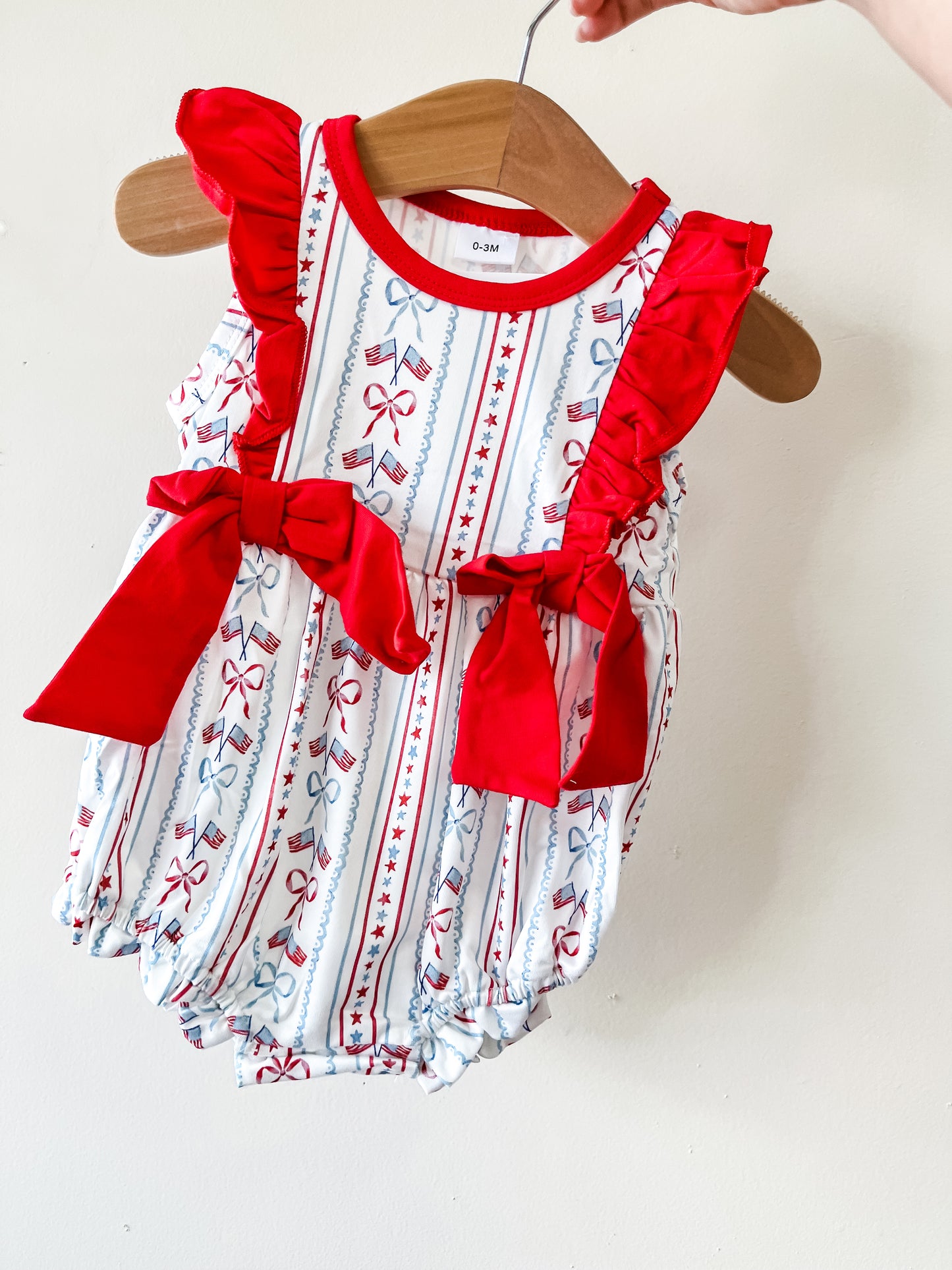 The Tiny Tot Bundles Patriotic Cherry Bubble features red ruffled straps with big bows and a white base adorned with red and blue stripes and bow patterns, shown hanging on a wooden hanger against a plain wall.