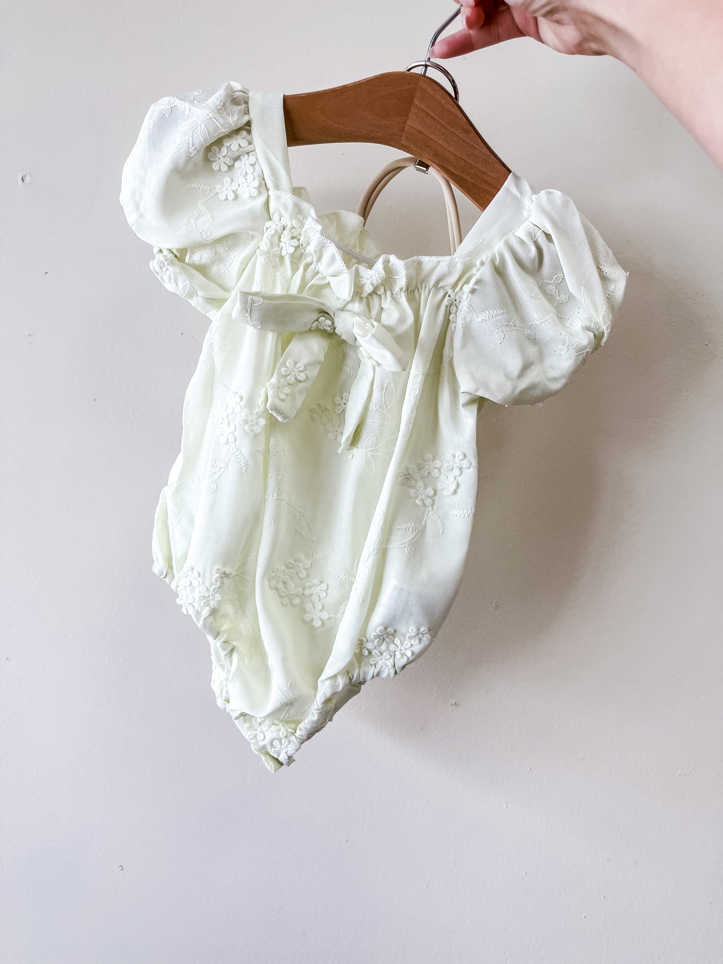 A hand holds Tiny Tot Bundles’ Dainty Bow Romper—a light yellow, short-sleeved baby outfit with floral embroidery, puff sleeves, and a neckline bow—against a plain light wall.