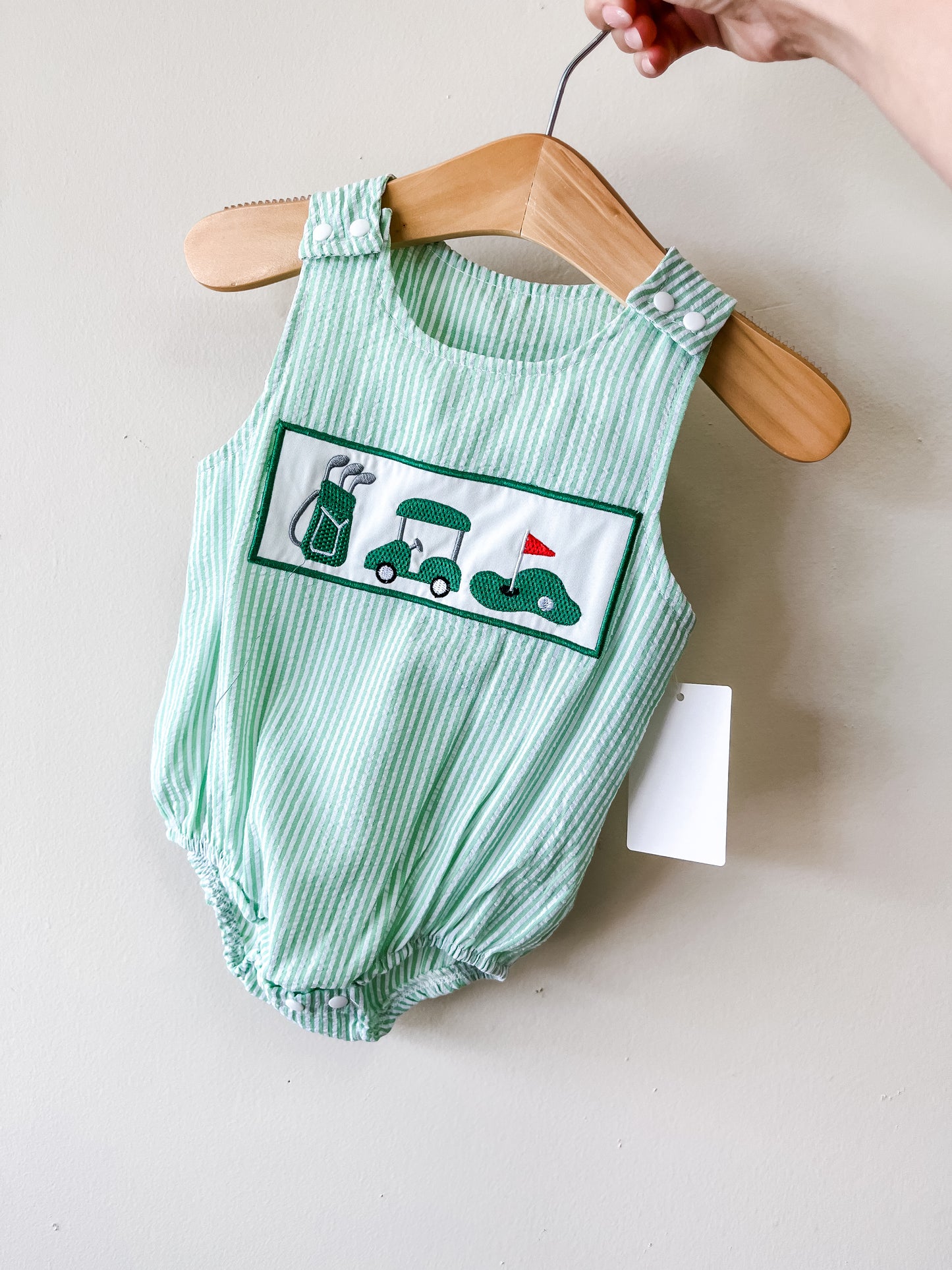 The Little Golfer Bubble by Tiny Tot Bundles is a green and white striped baby romper with embroidered golf-themed designs—golf bag, golf cart, and putting green with red flag—displayed on a hanger with a white tag.