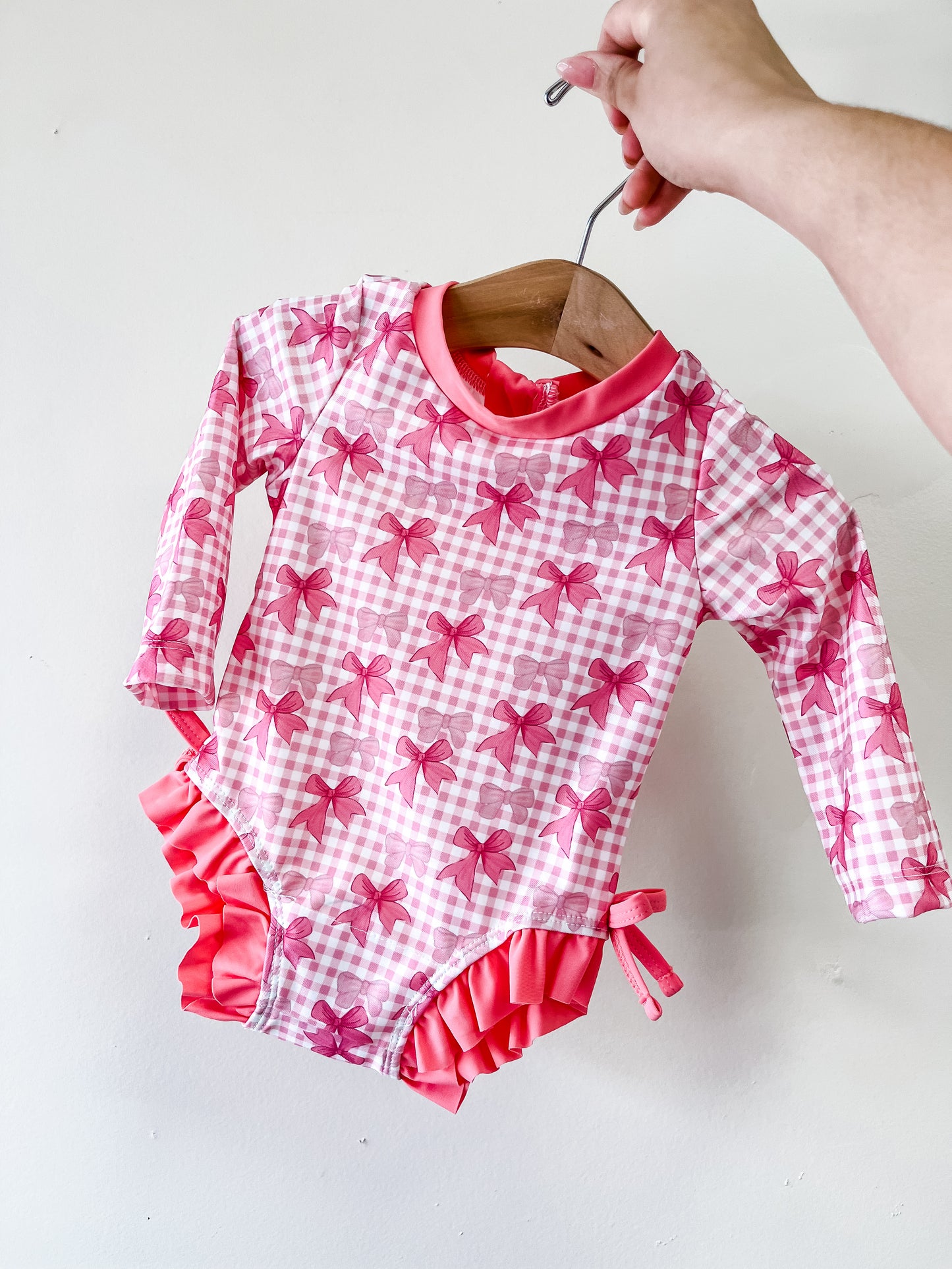 A hand holds the Pretty Bow Babe Swimwear by Tiny Tot Bundles, a pink and white checkered long-sleeve baby swimsuit with pink bows and ruffled leg trim, displayed on a wooden hanger against a white background.