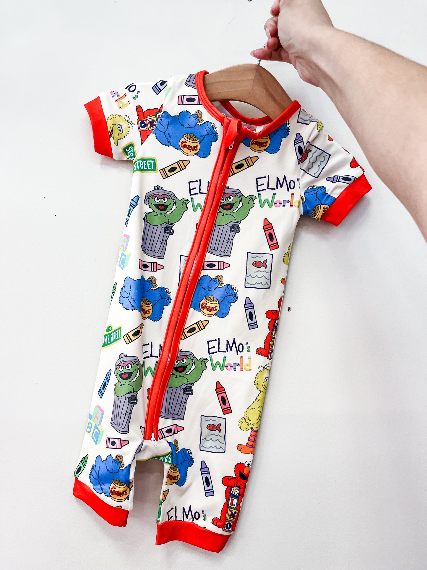 A hand holds up the Sesame Street Shorty by Tiny Tot Bundles, a baby romper featuring Elmo, Oscar the Grouch, Cookie Monster, Big Bird, colorful crayons, “Elmos World” text, red trim, and a convenient zipper.