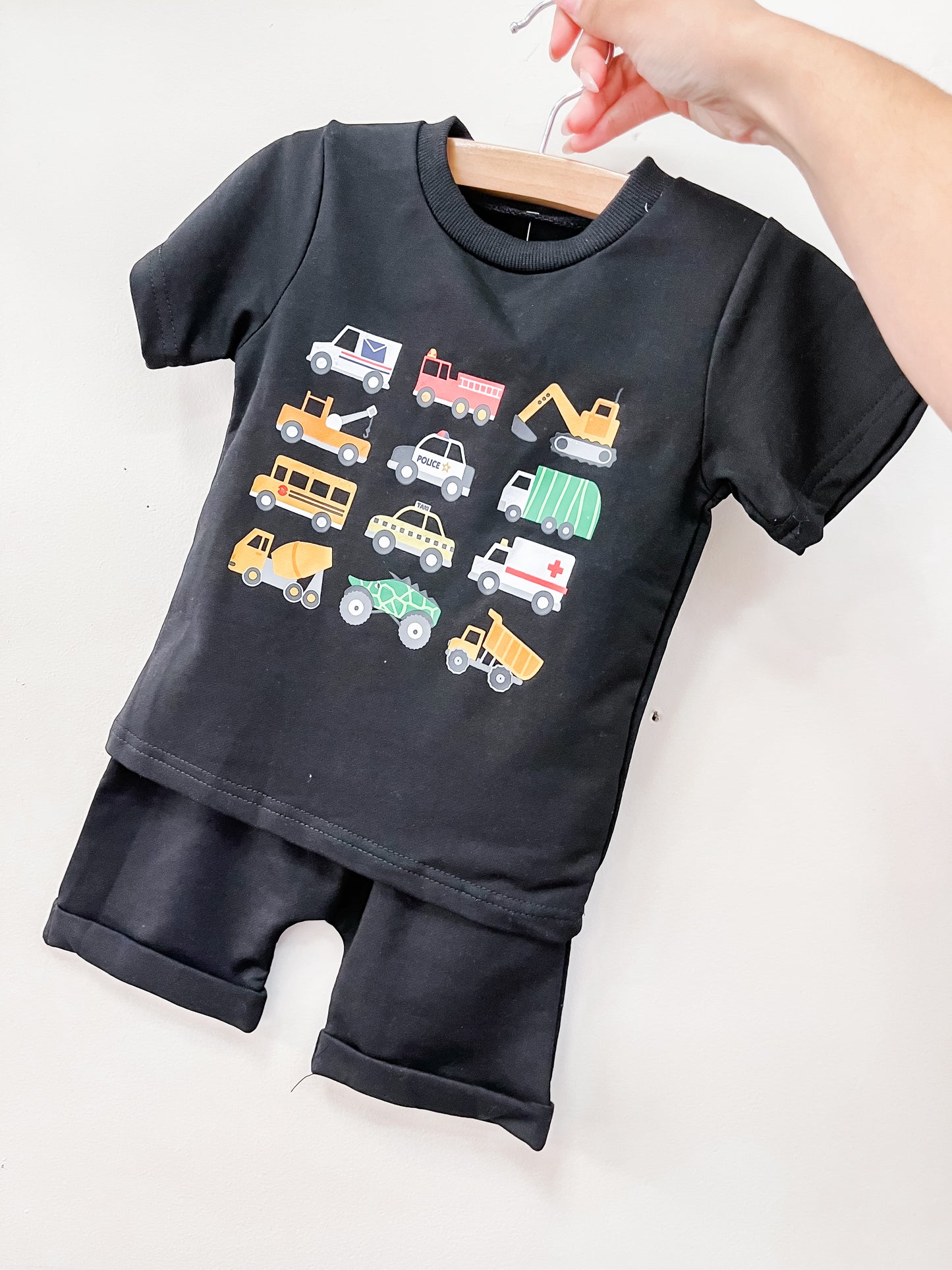 The Tiny Tot Bundles Transportation Crew Shorts Set is a black short-sleeve toddler outfit with matching shorts, featuring colorful vehicle illustrations like cars, trucks, buses, and construction equipment on the shirt. Displayed on a hanger.