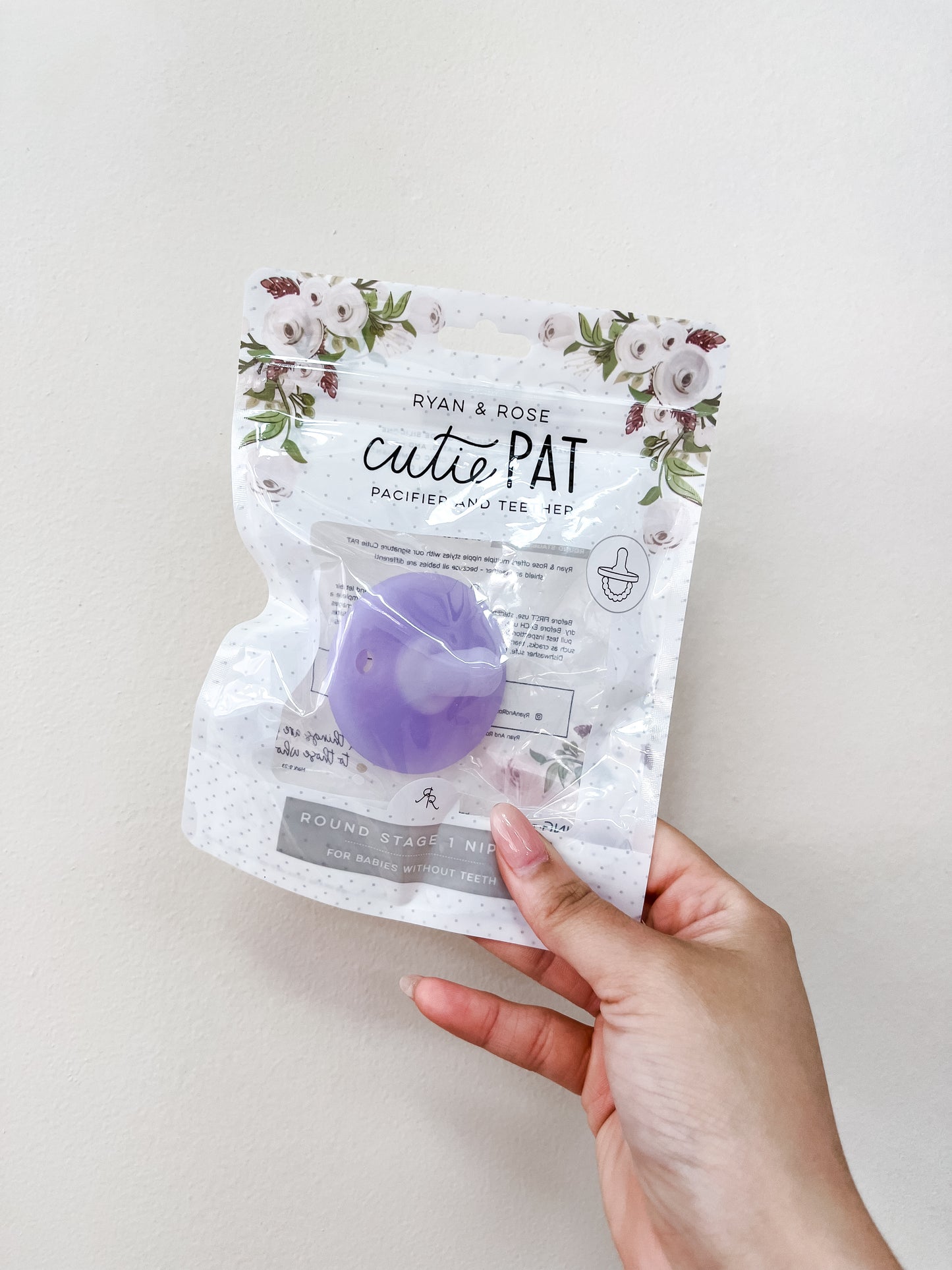 A hand holds a packaged ryan & Rose Cutie Pat Pacifier - Clear Purple against a plain light background. The packaging features floral designs at the top.