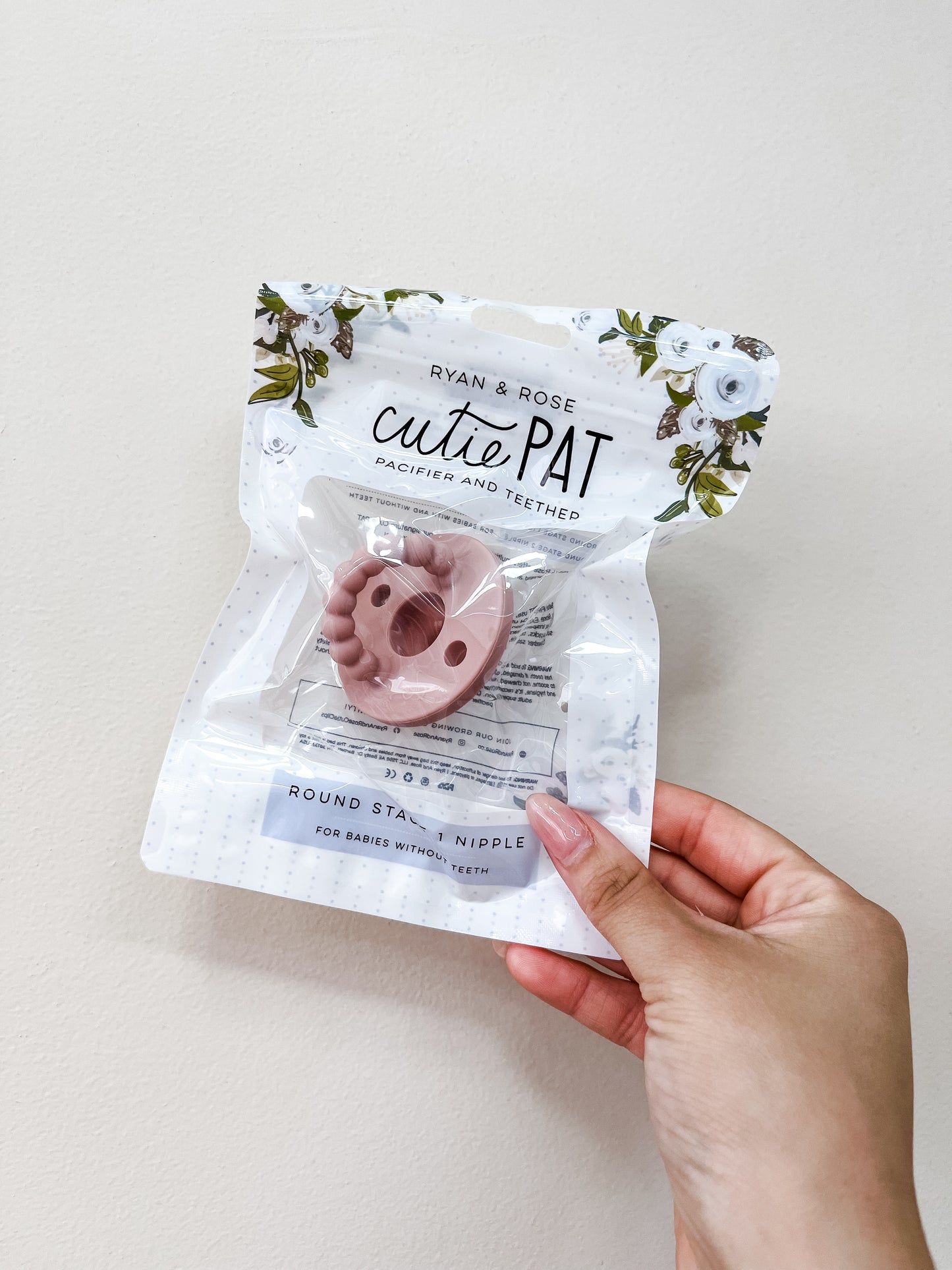 A hand holds a clear package labeled ryan & Rose, containing the Cutie Pat Pacifier - Muave with floral designs on the packaging.