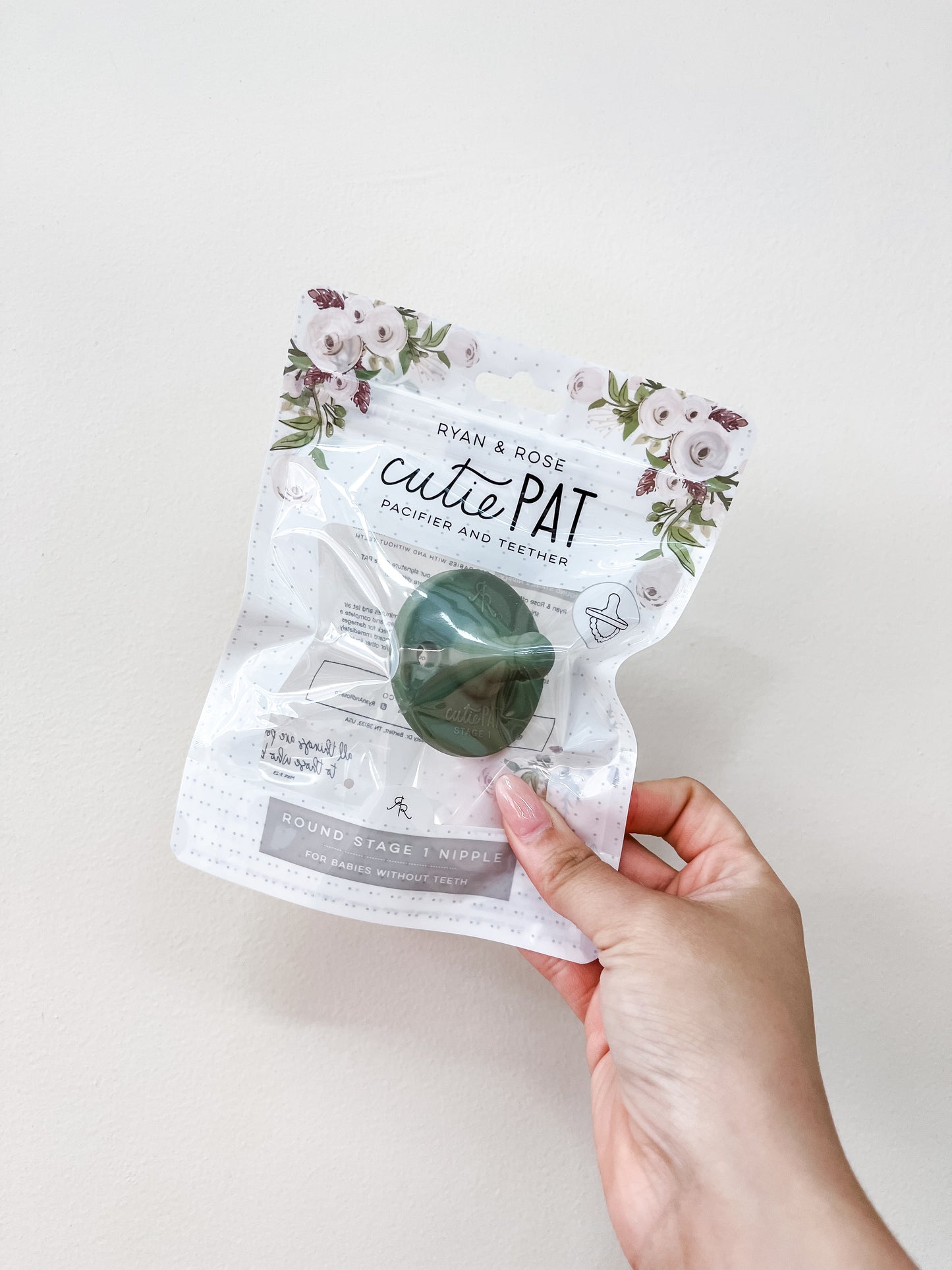 A hand holds a clear package of the ryan & Rose Cutie Pat Pacifier in Sage green, showcasing the floral design and green pacifier inside.