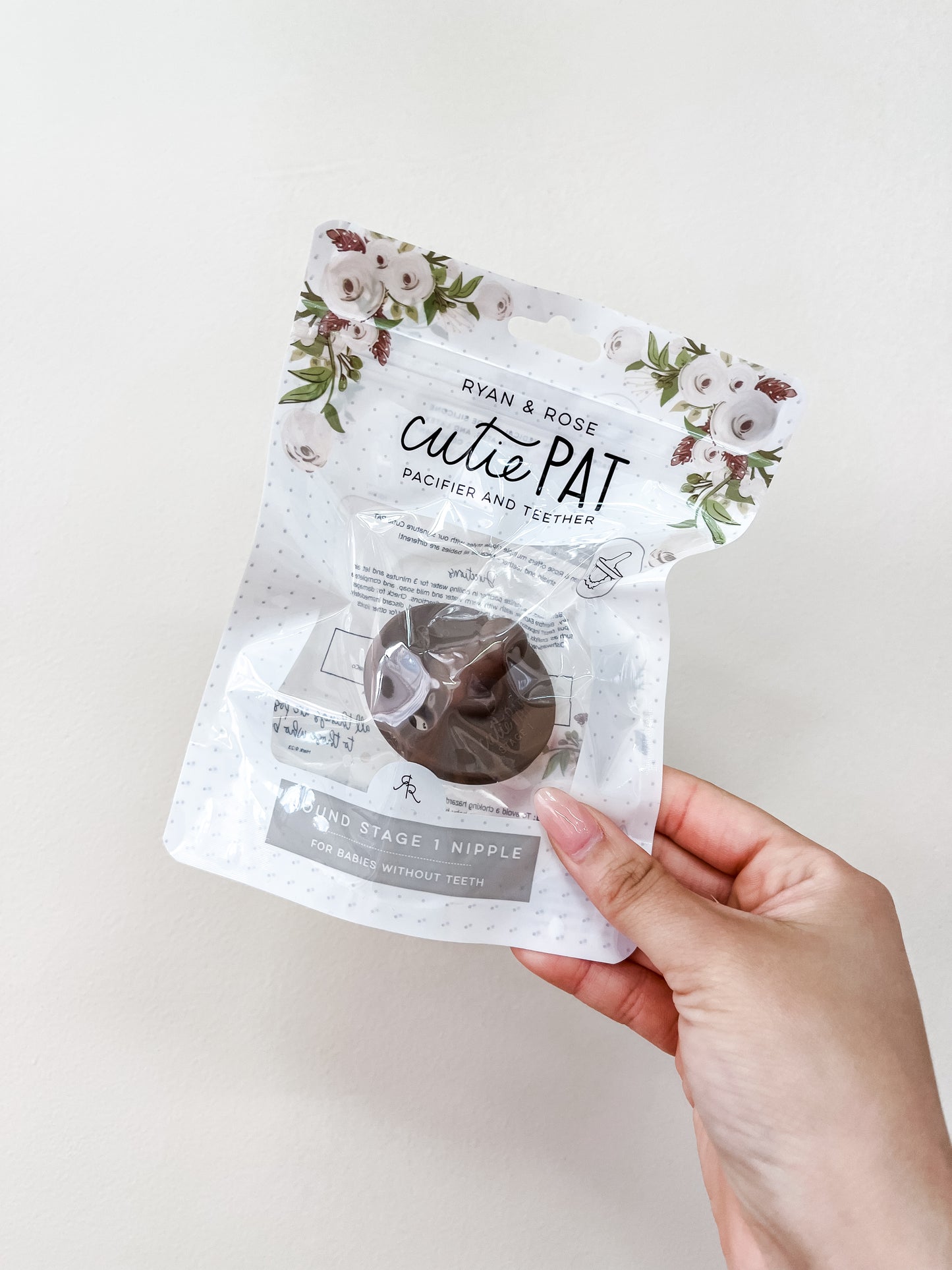 A hand holds a packaged Cutie Pat Pacifier - Brown by ryan & Rose, featuring floral design at the top of the packaging, against a plain light background.