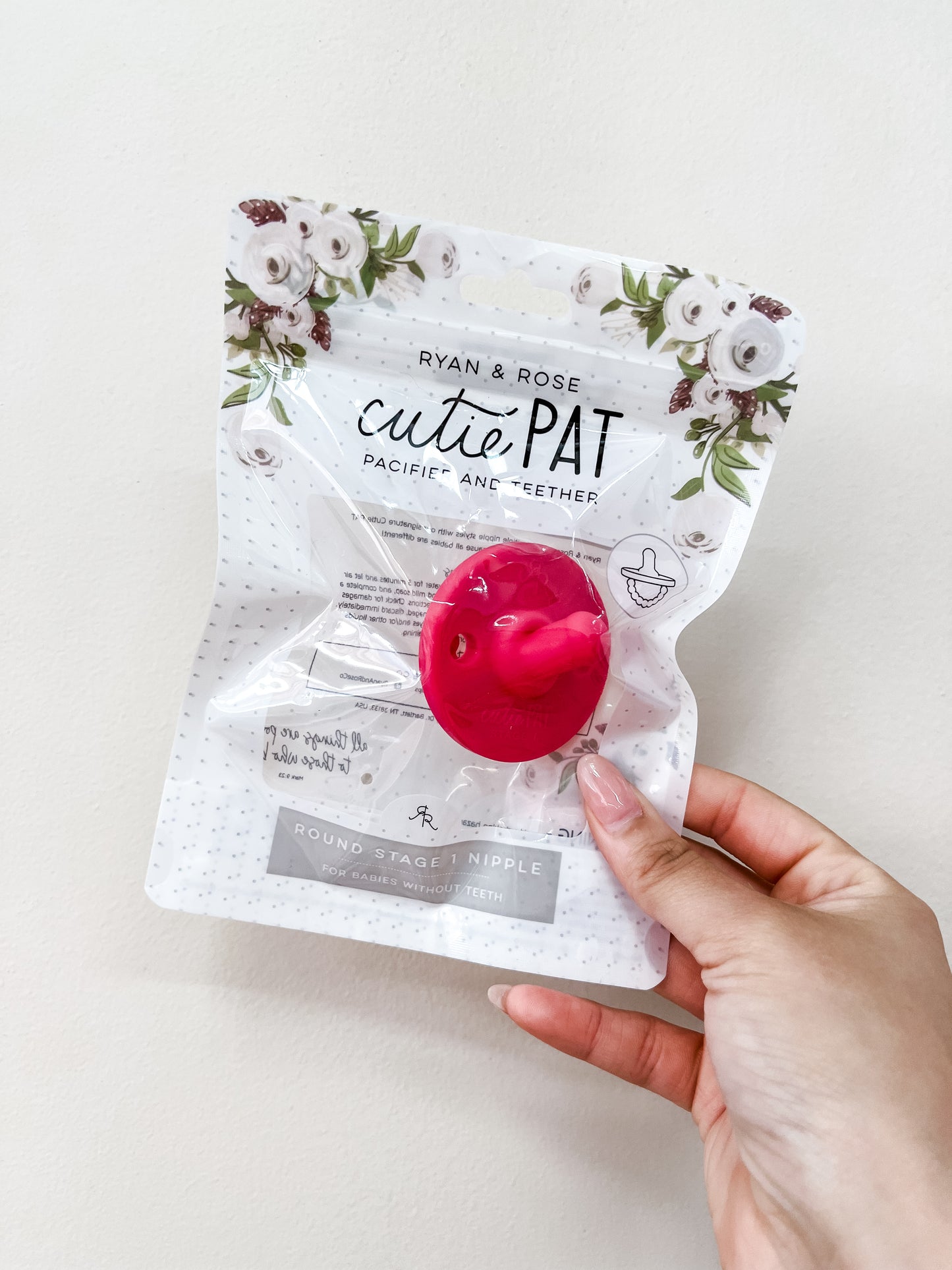 A hand holds a ryan & Rose Cutie Pat Pacifier - Hot Pink, visible in its floral-topped package against a plain white background.