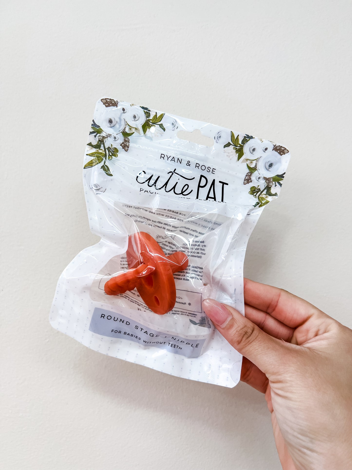 A hand holds a packaged ryan & Rose Cutie Pat Pacifier - Orange. The clear packaging features floral accents and product details on the label.